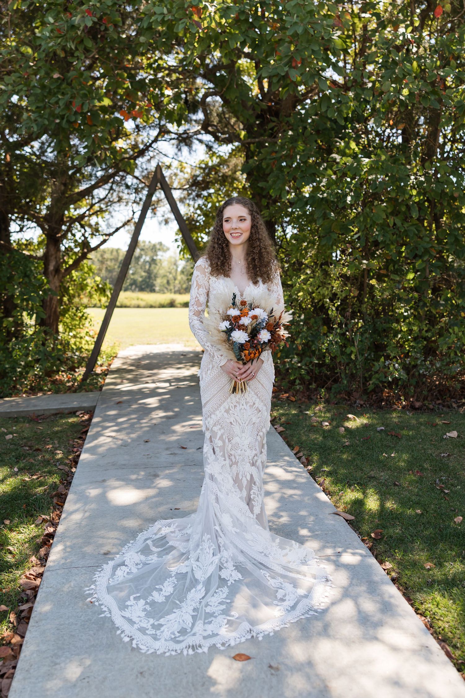 A bride in a lace wedding dress poses outdoors in dappled sunlight holding a colorful autumn bouquet.