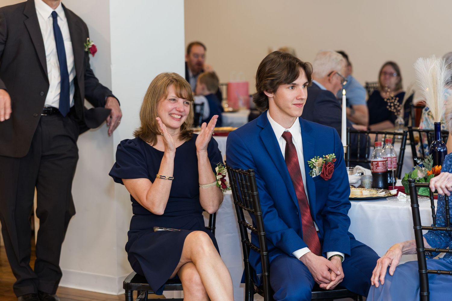 Woman in navy dress claps while young man in blue suit sits beside her at a wedding reception.