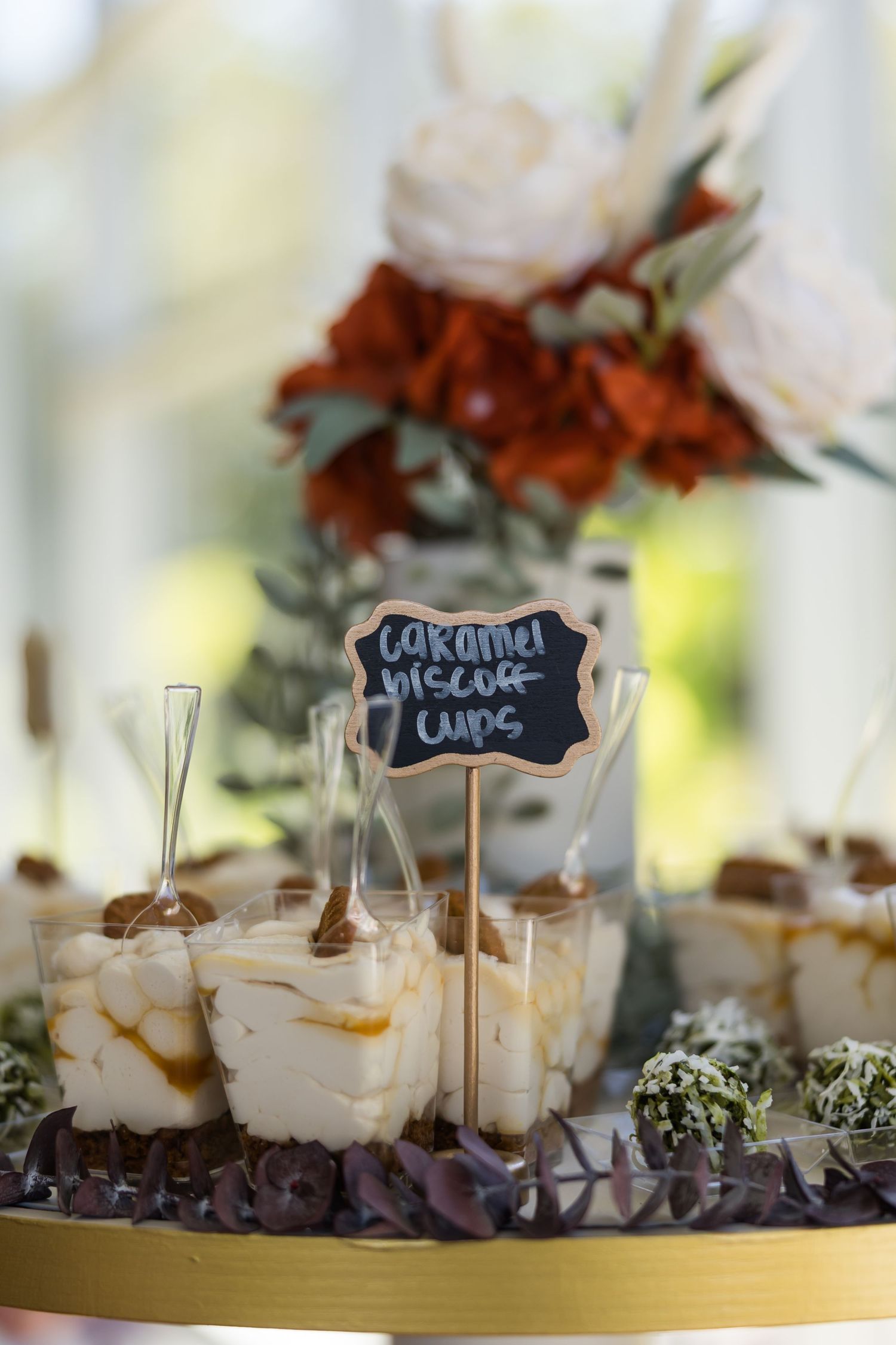 Caramel biscoff cups labeled with chalkboard sign on gold dessert table with floral centerpiece.