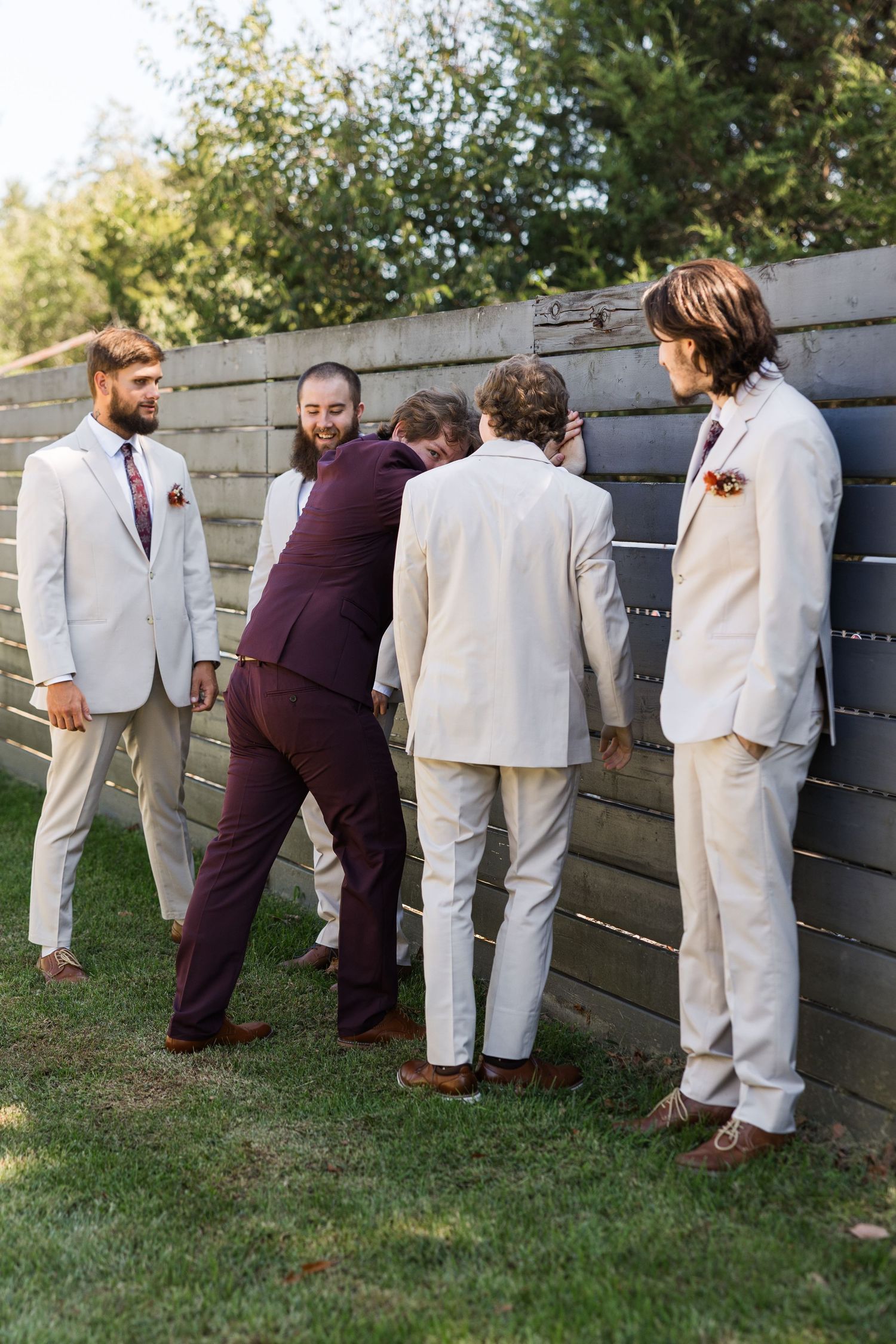 Groom in burgundy suit joking with groomsmen in tan suits near a wooden fence at outdoor wedding venue.