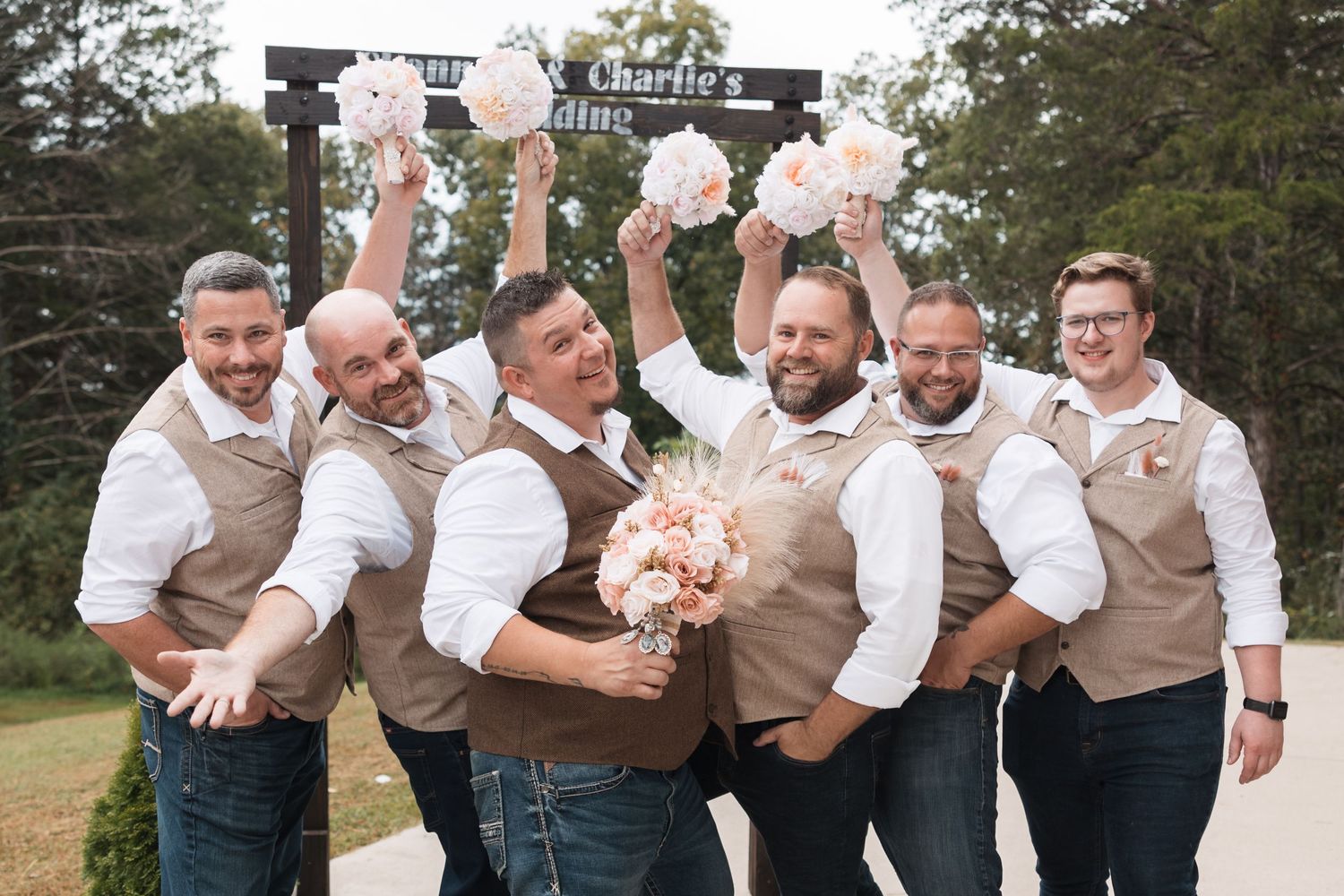 Groomsmen in tan vests and jeans celebrate with bouquets at an outdoor wedding venue sign.