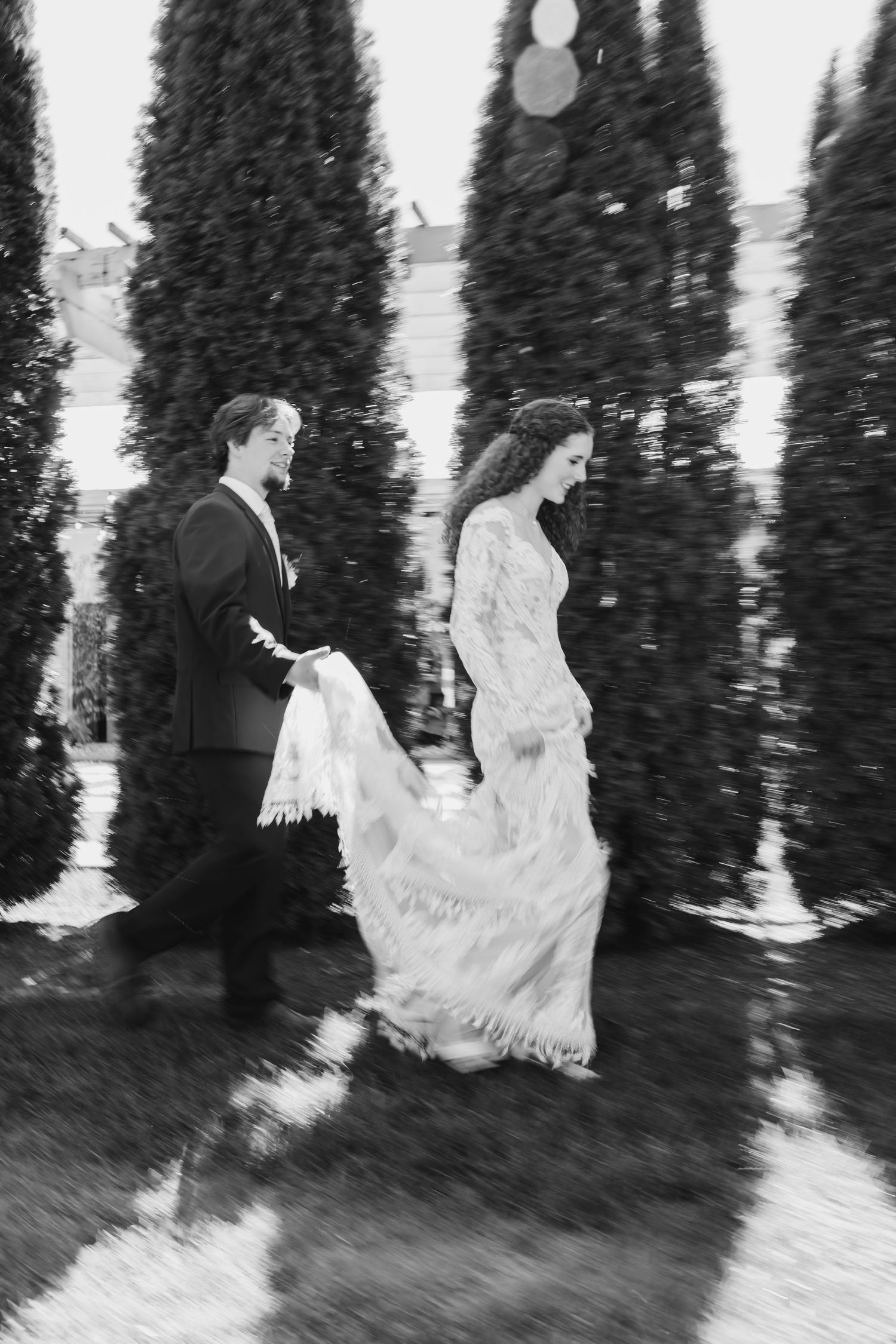 Black and white photo of a bride in lace gown and groom in suit walking among tall cypress trees.
