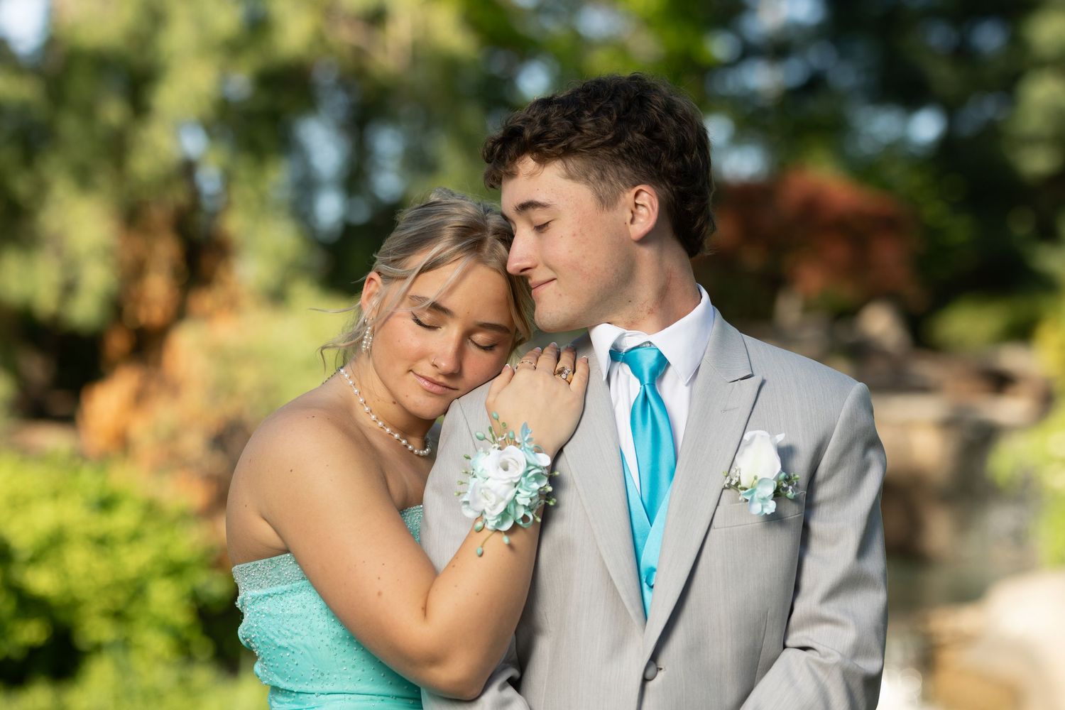 A couple in prom attire, she in mint dress with corsage, he in gray suit with teal tie, pose outdoors.