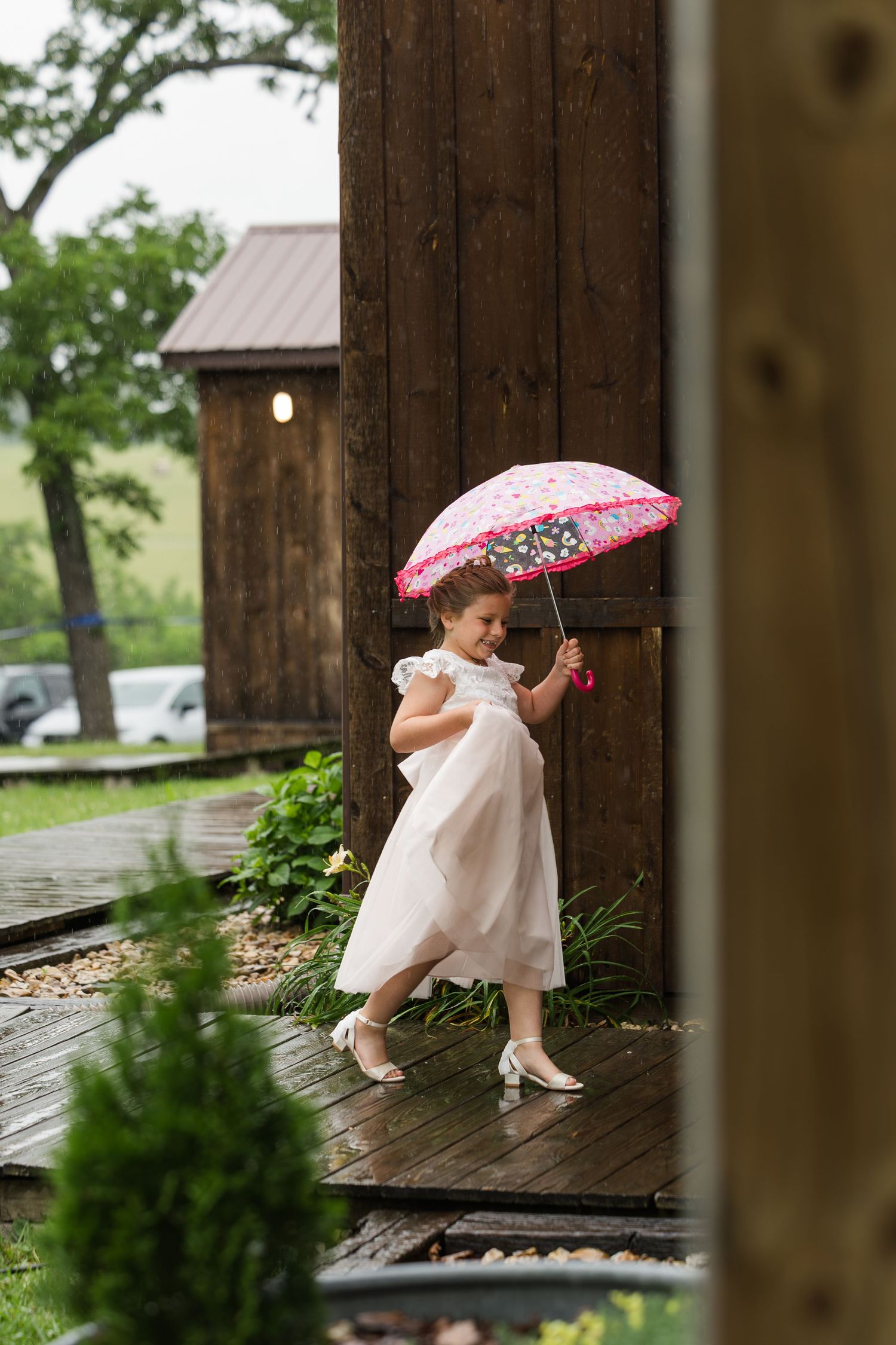Young girl in white dress holding pink umbrella on wooden deck near rustic barn during rainy day.
