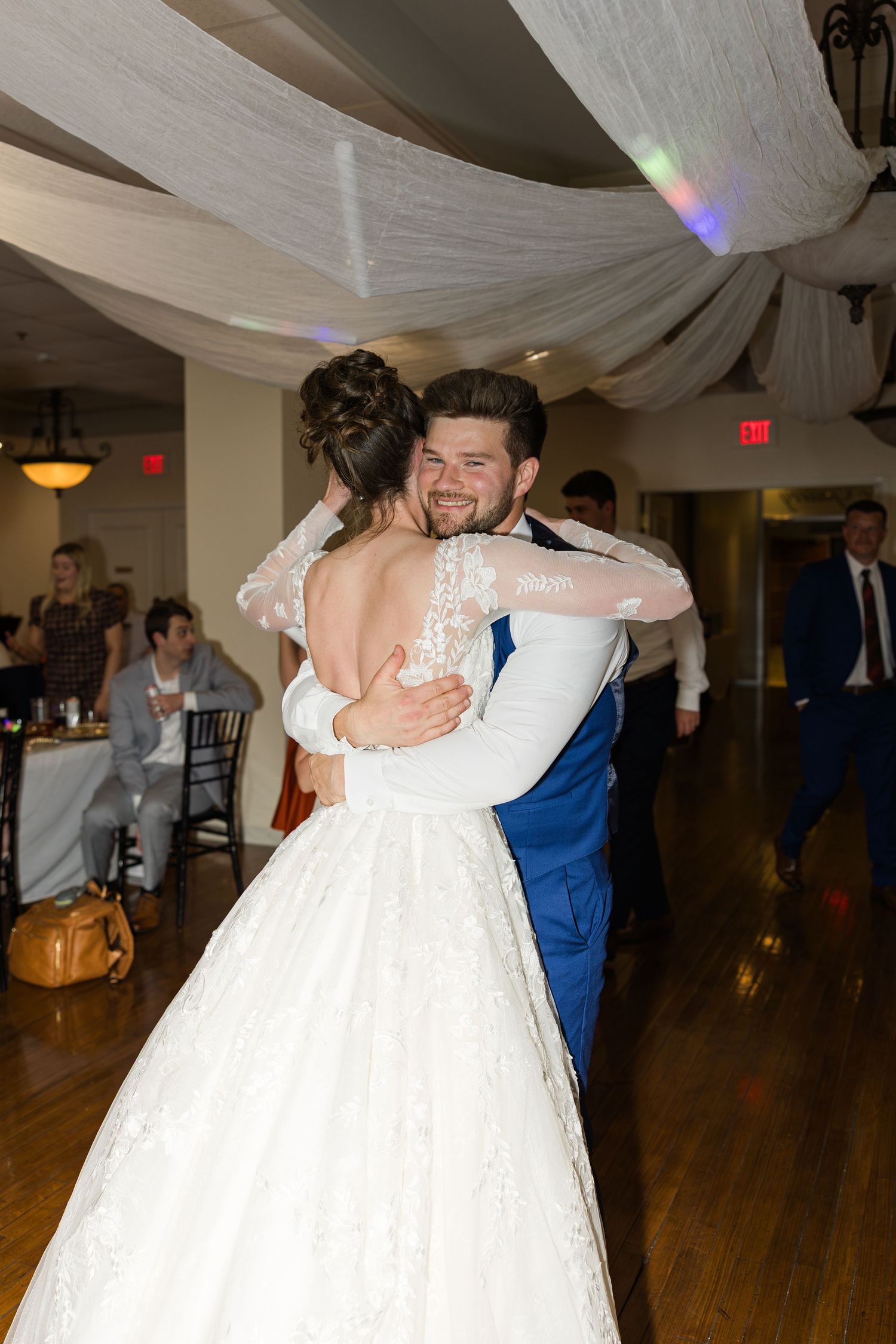 Groom smiles joyfully while sharing first dance with bride in lace gown at wedding reception.