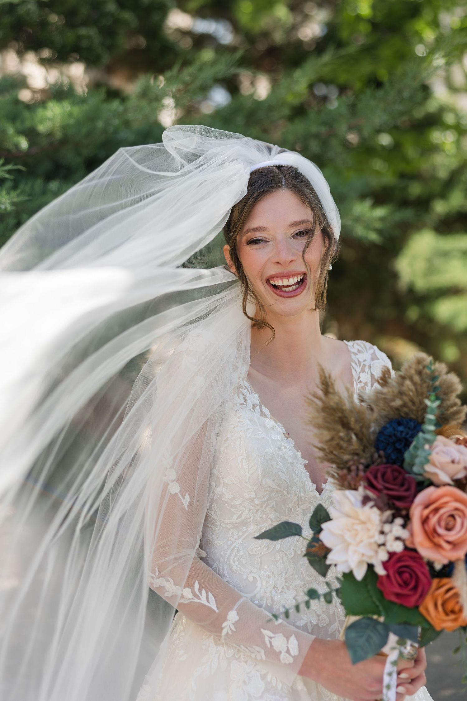 Joyful bride in lace wedding dress laughs as her veil flows in the wind while holding a colorful bouquet.