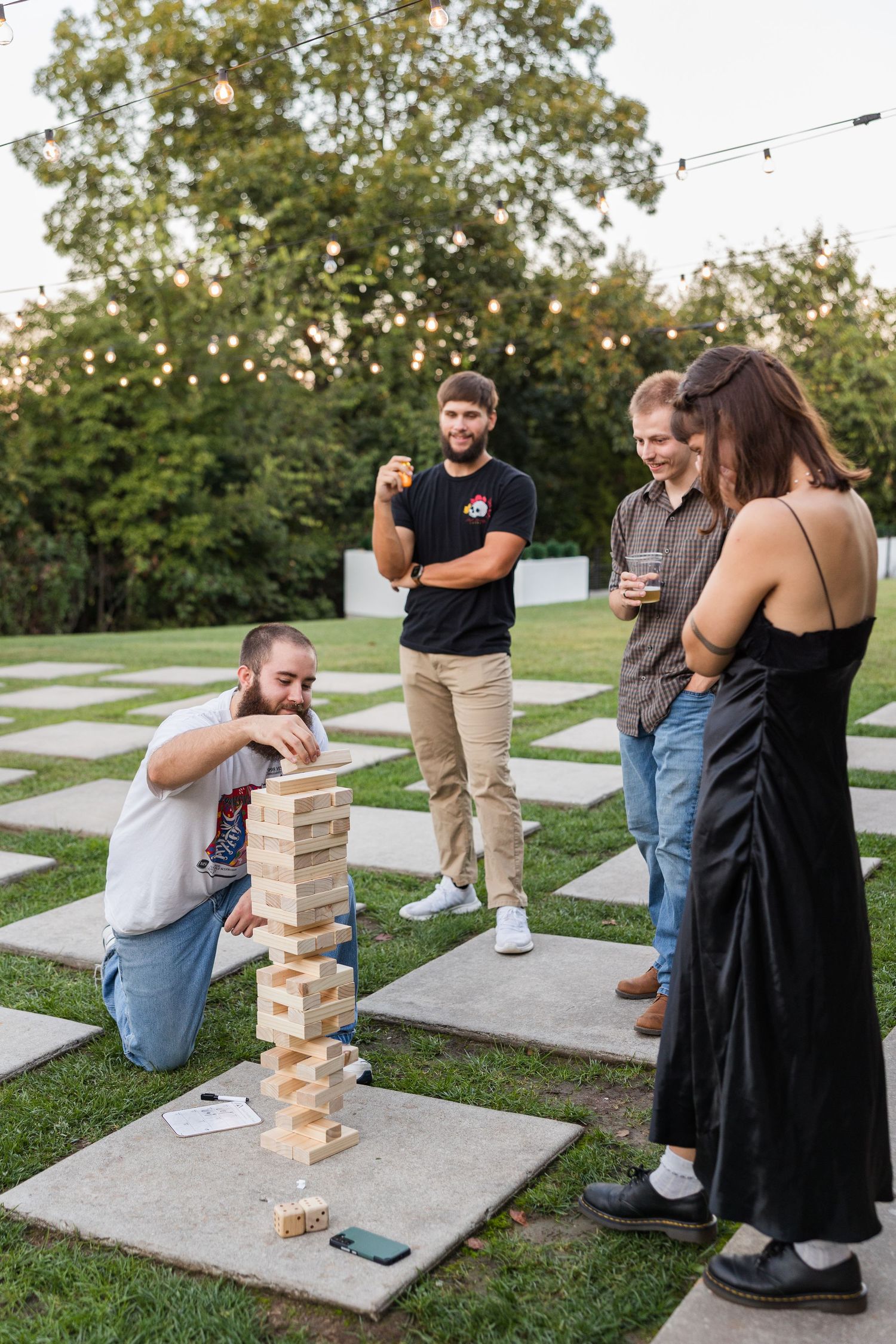 Wedding guests playing giant Jenga on a stone patio at an outdoor evening reception with string lights.