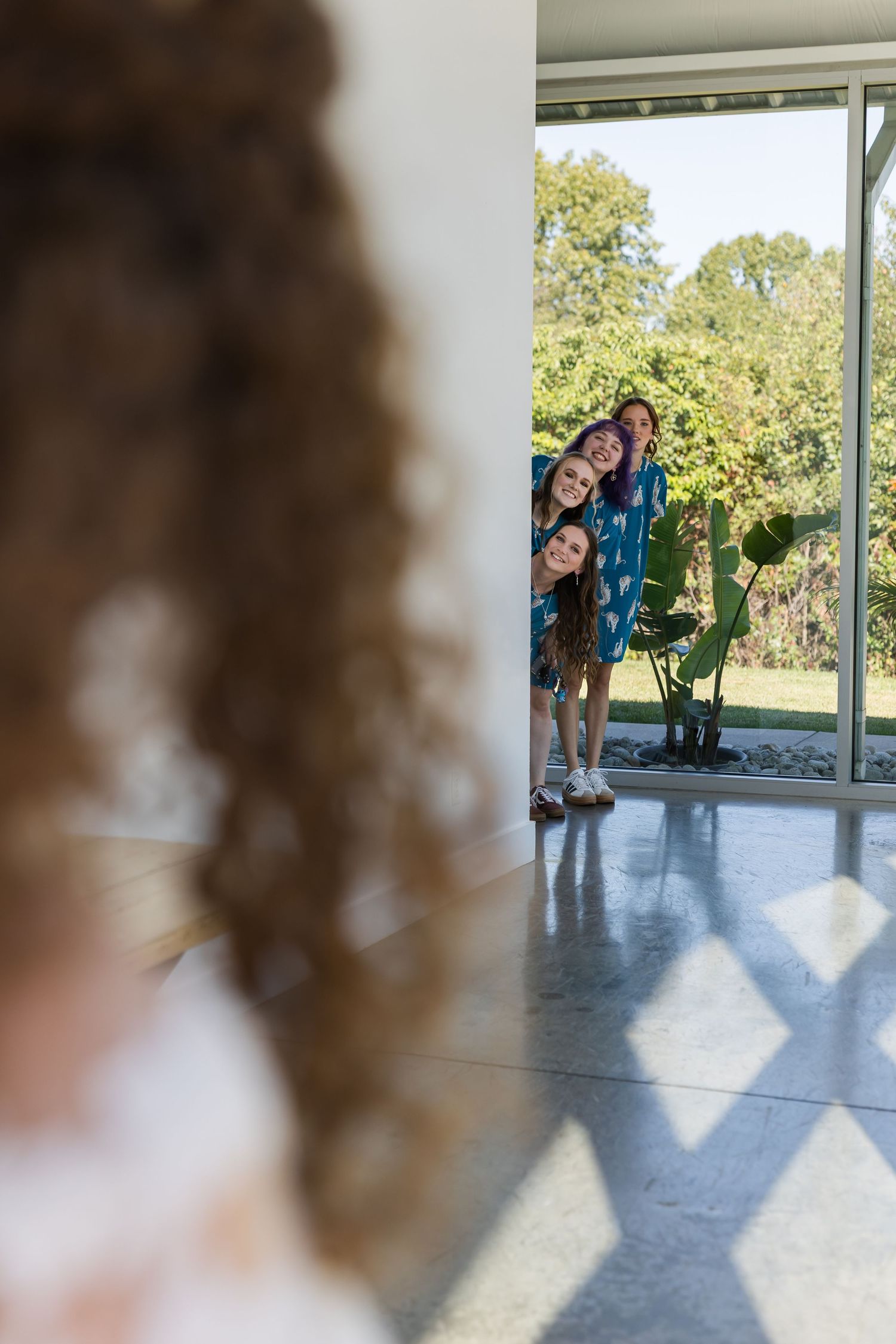 A group of smiling people in teal outfits peeking around a corner inside a modern home with large windows.