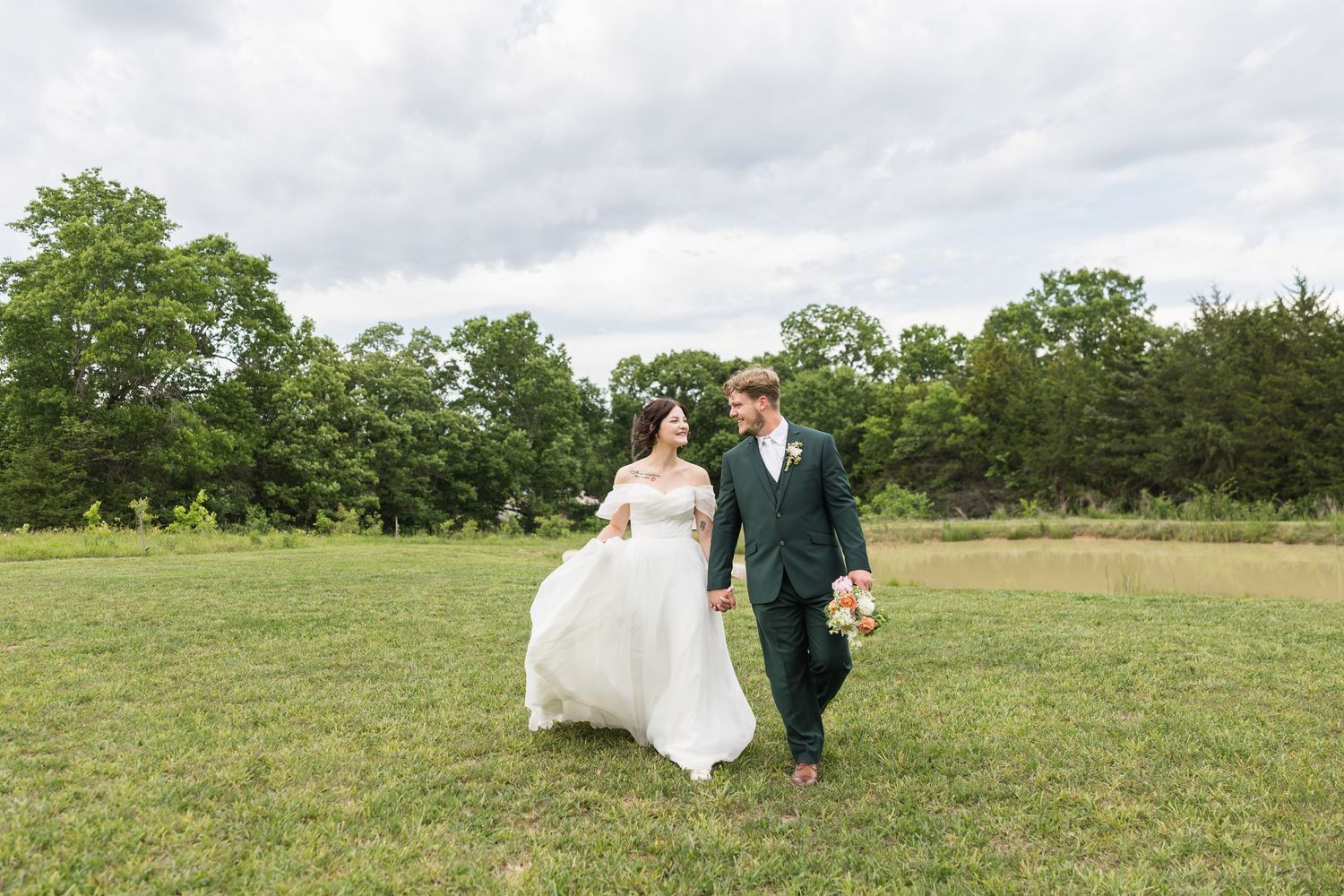 Bride and groom walking hand in hand through green meadow on cloudy wedding day.