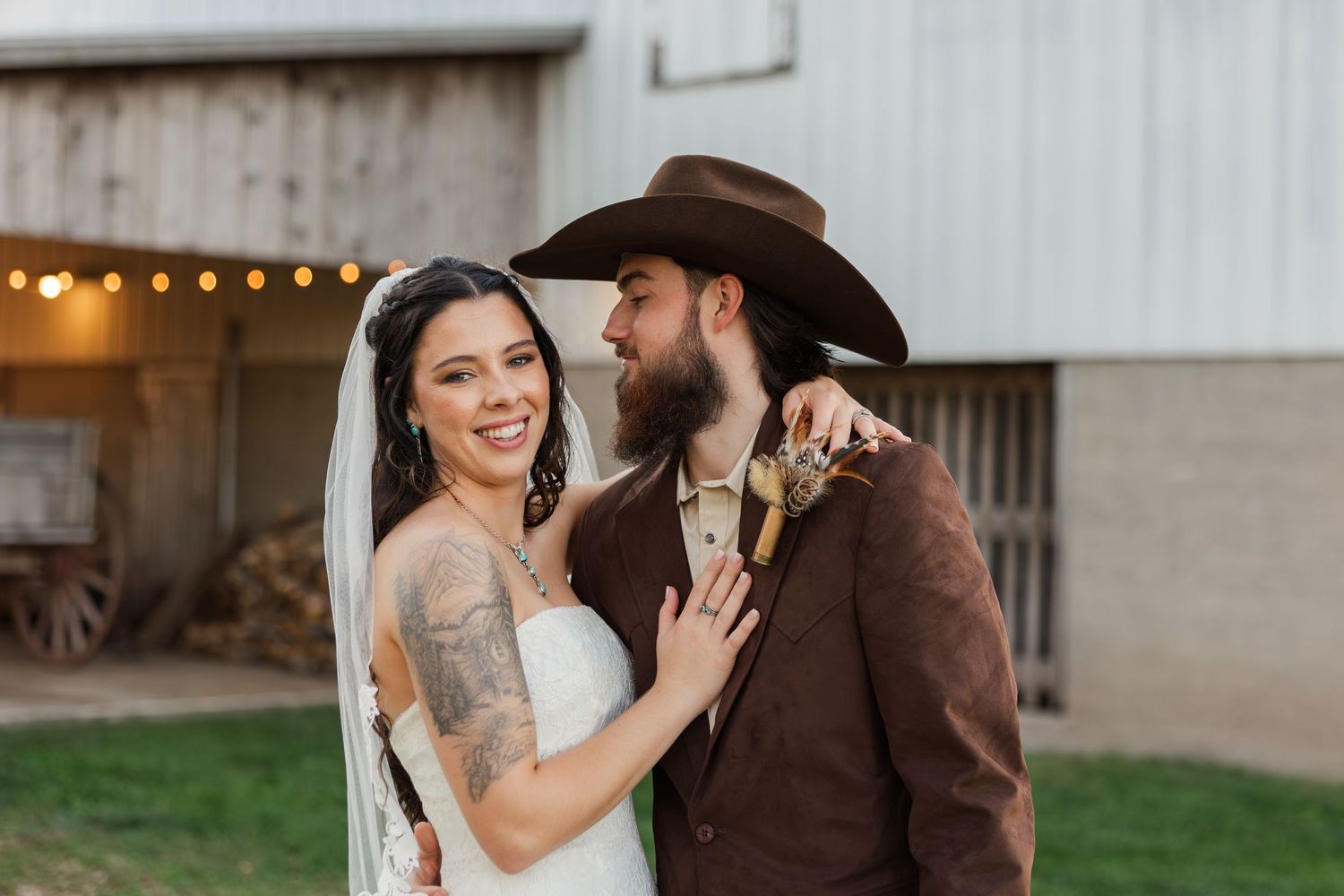 Bride in white dress and veil smiles beside groom in brown suit and cowboy hat at rustic barn wedding.