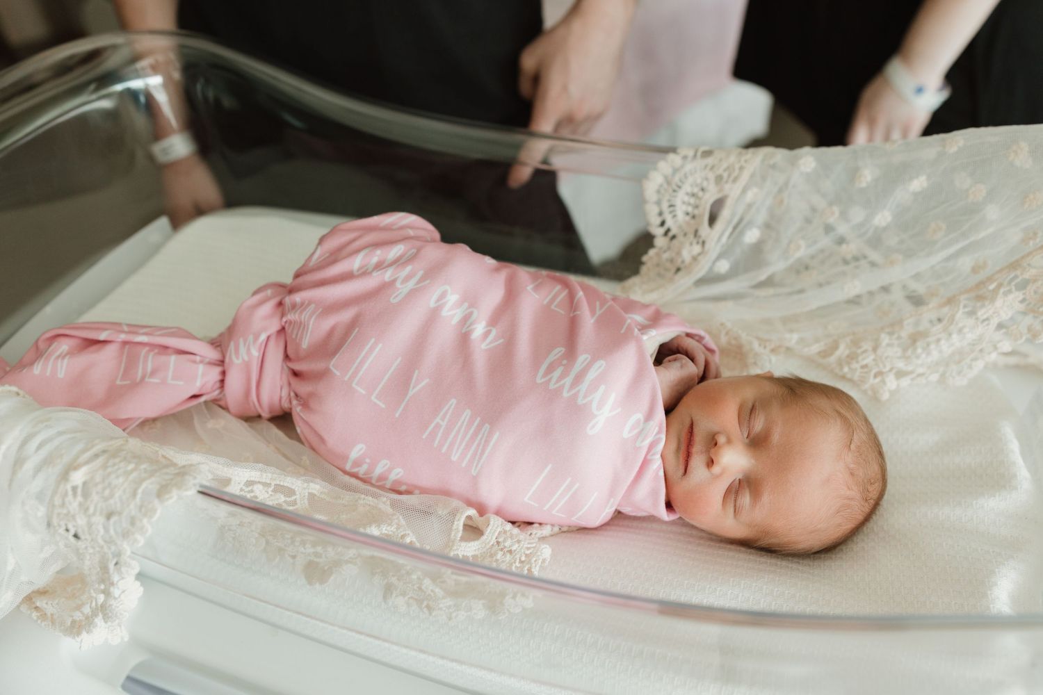 Newborn baby in pink sleepwear rests peacefully on white lace blanket during professional photography session.