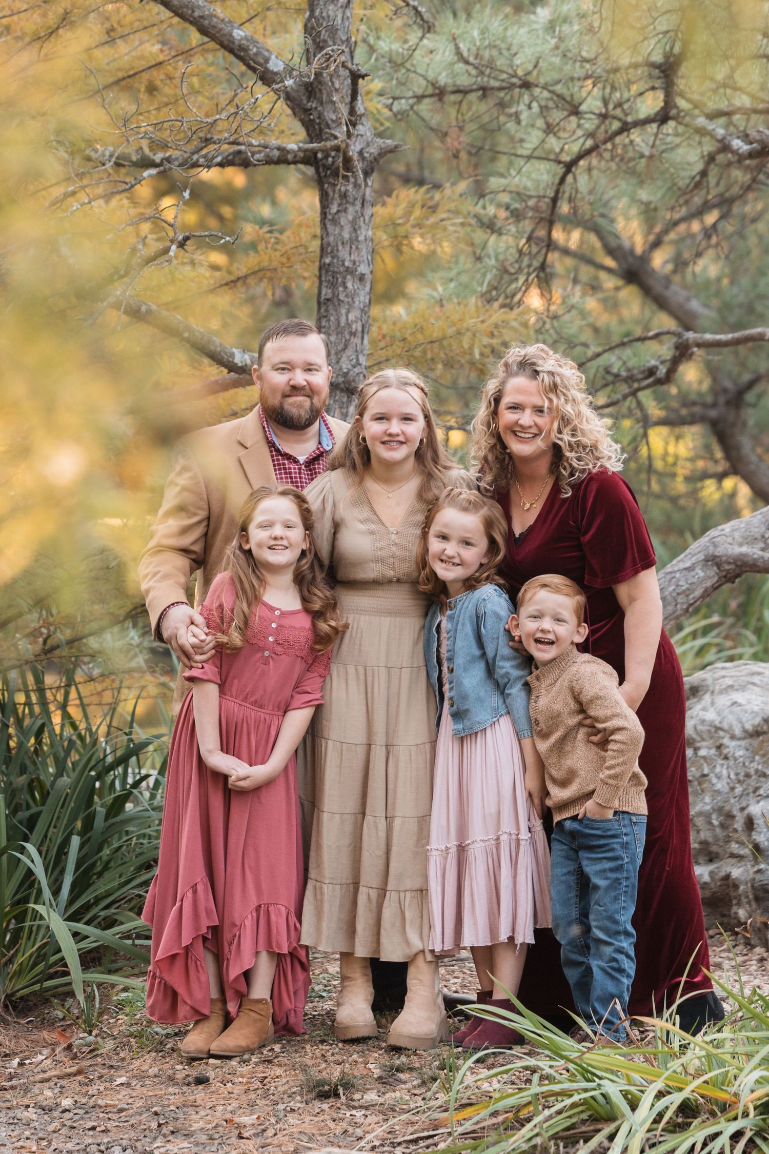 Family of six poses together outdoors wearing coordinated fall colors against autumn foliage.