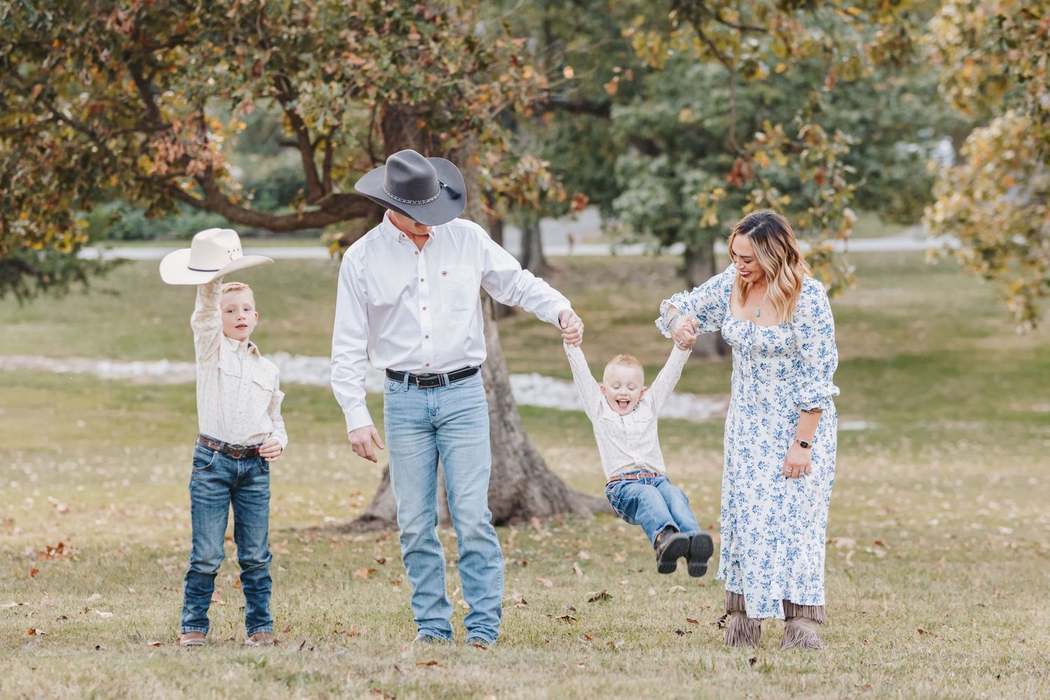 A playful family holds hands and swings together in a pastoral park setting.
