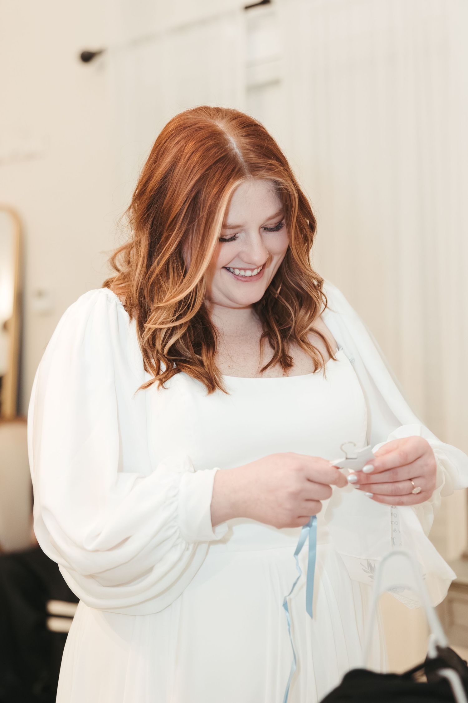 A bride in a white long-sleeved dress smiles while holding something in her hands.