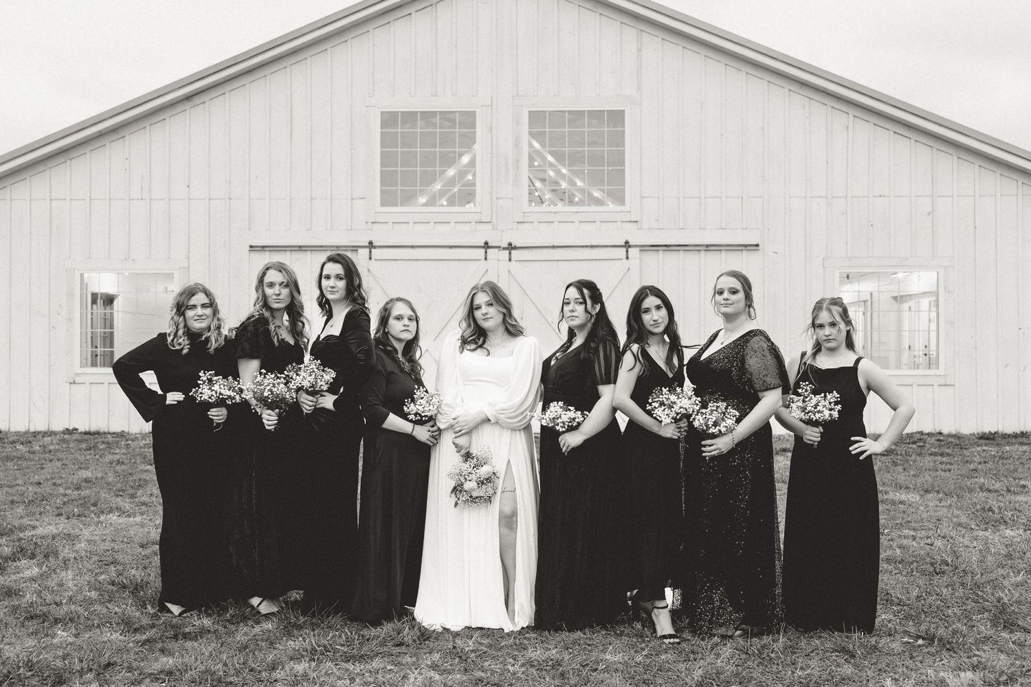 Wedding party wearing black and white dresses pose in front of a rustic white barn with arched windows.
