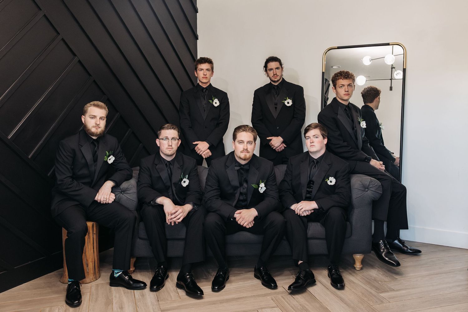 A group of groomsmen in black suits pose together in a modern room with a black accent wall and mirror.