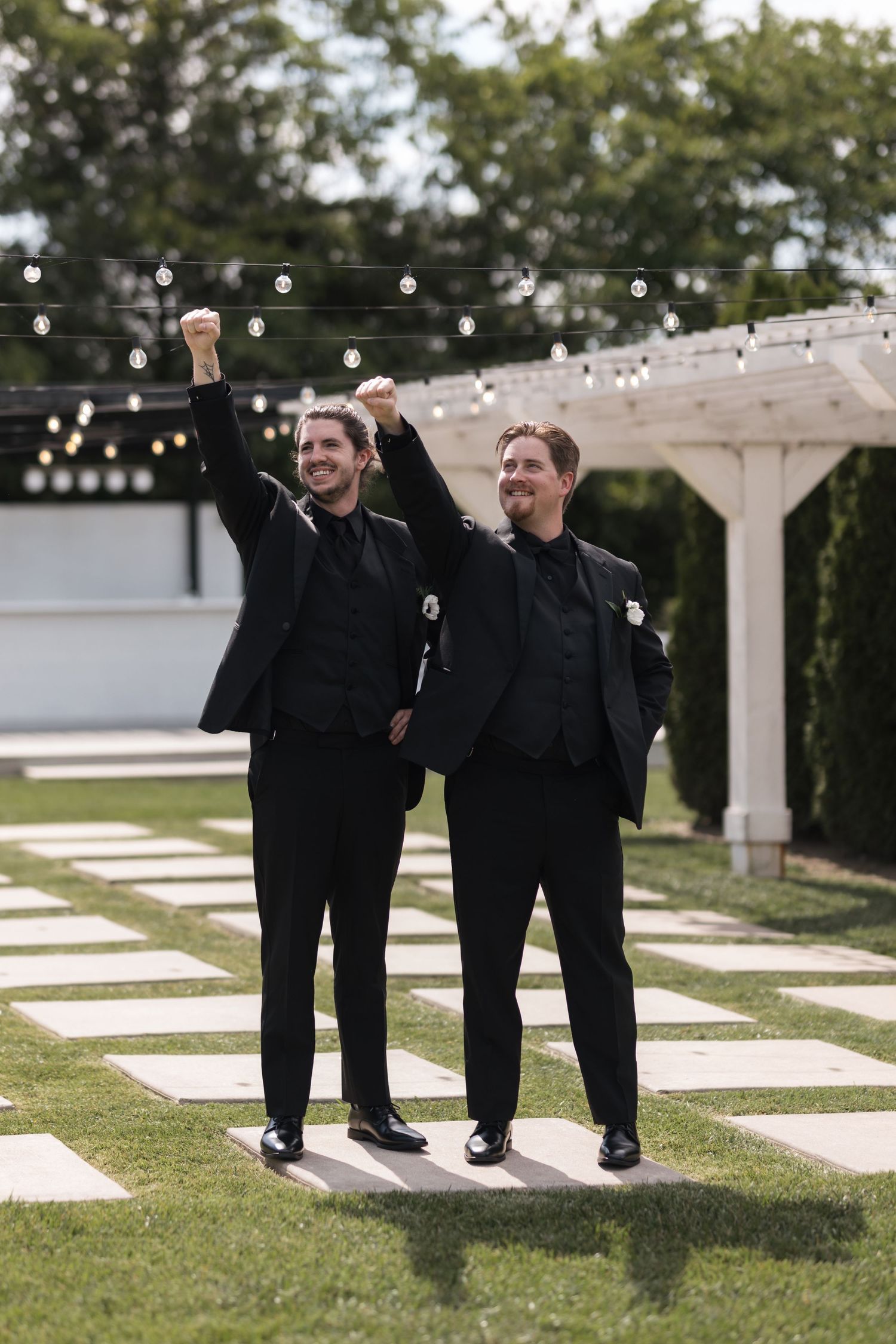 Two people in black suits celebrate with raised arms under string lights at outdoor venue.