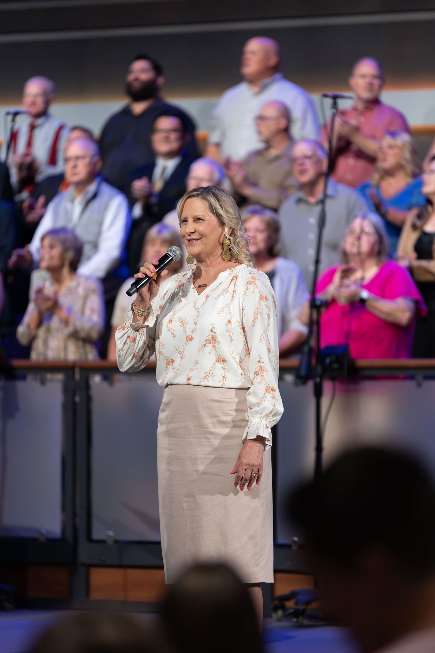 Performer in cream blouse and skirt sings on stage with choir behind.