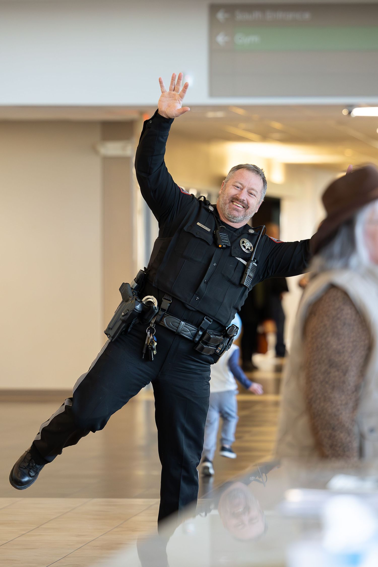 Police officer in uniform strikes an enthusiastic pose while waving in a hallway.