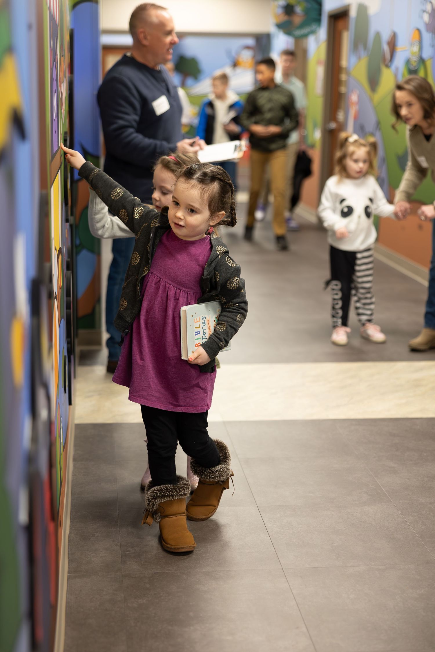 Small child in purple dress exploring interactive wall display in school hallway.