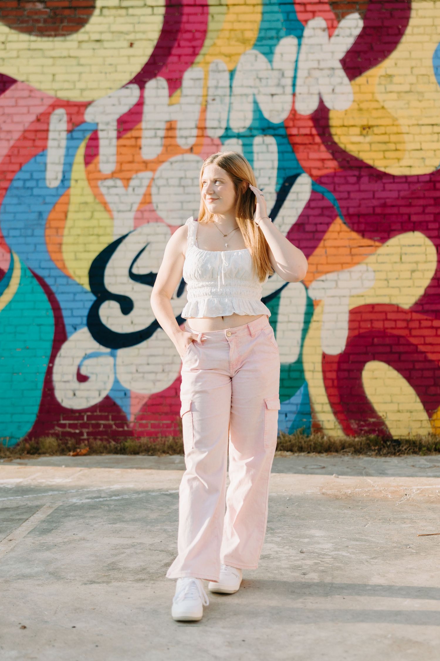 A person poses against a vibrant graffiti wall wearing a white cropped top and pink cargo pants with white sneakers.