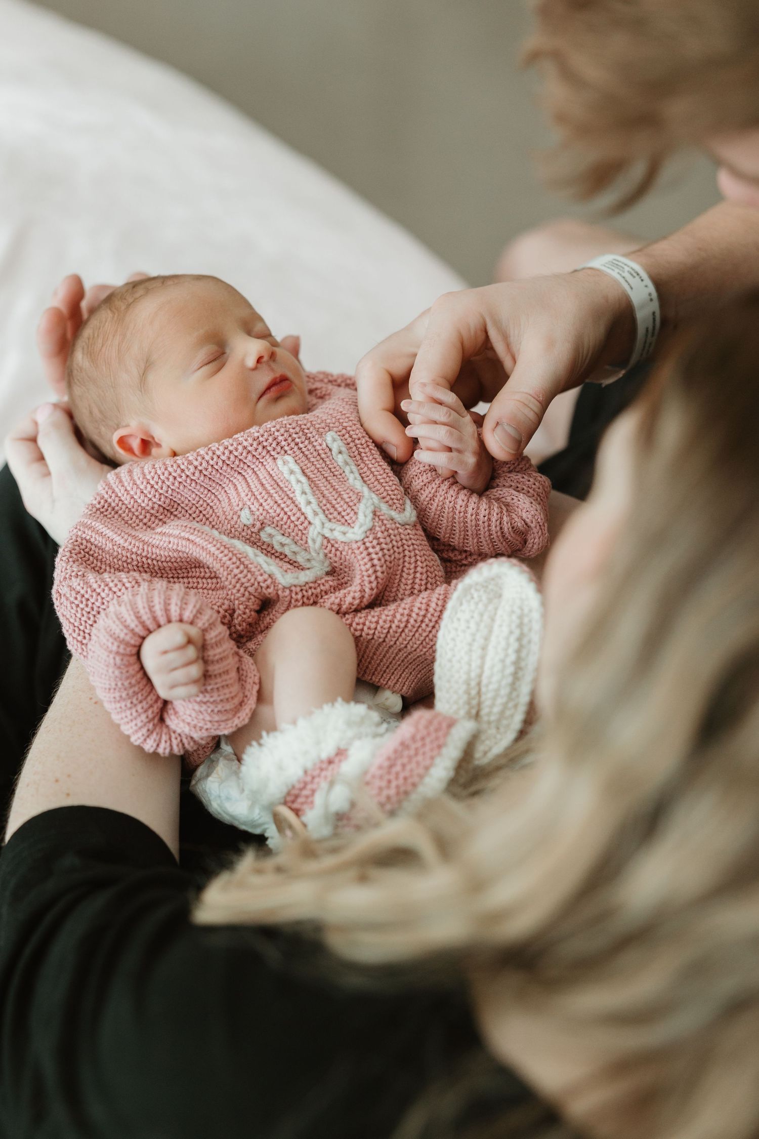 A newborn infant wearing a pink knit sweater rests on someone's lap during a close-up shot.