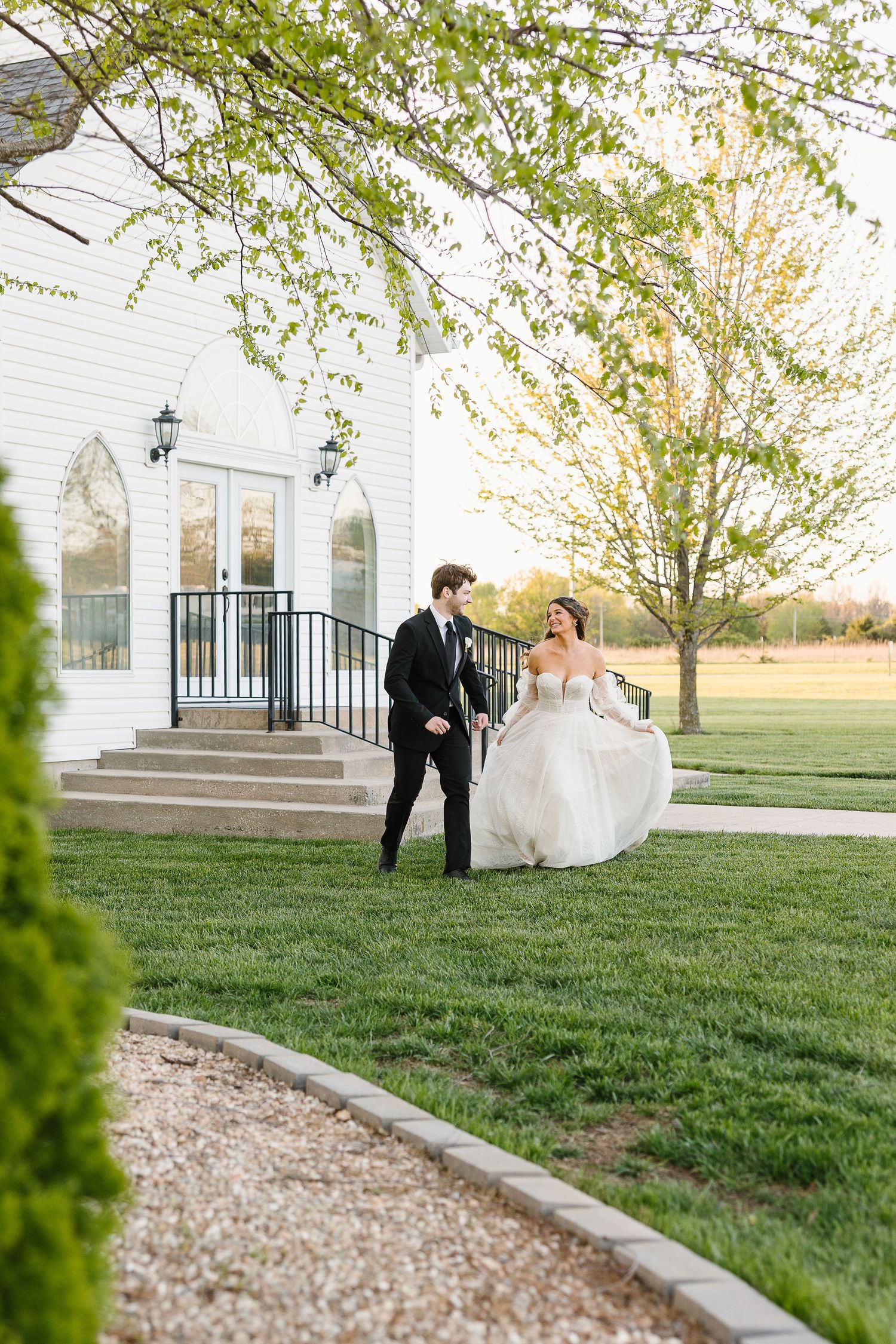 A bride and groom share a joyful moment outside a white chapel with arched windows and gravel pathways.