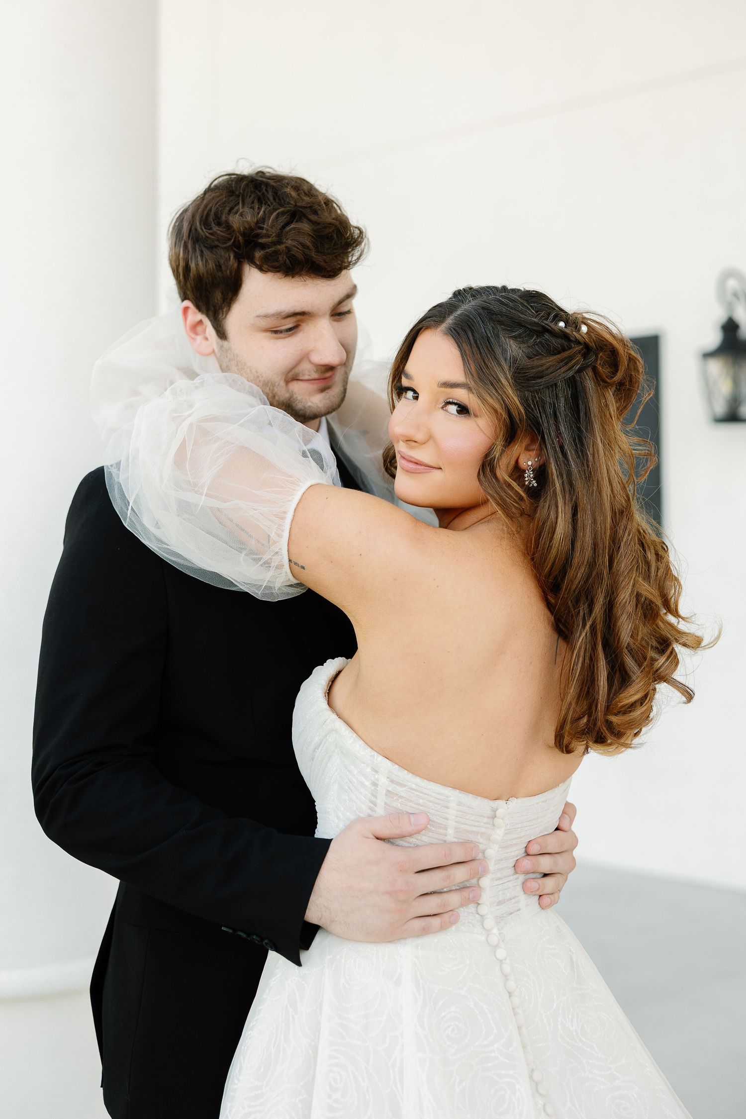 A romantic wedding portrait of a couple embracing, with the bride in a white off-shoulder gown looking over her shoulder.