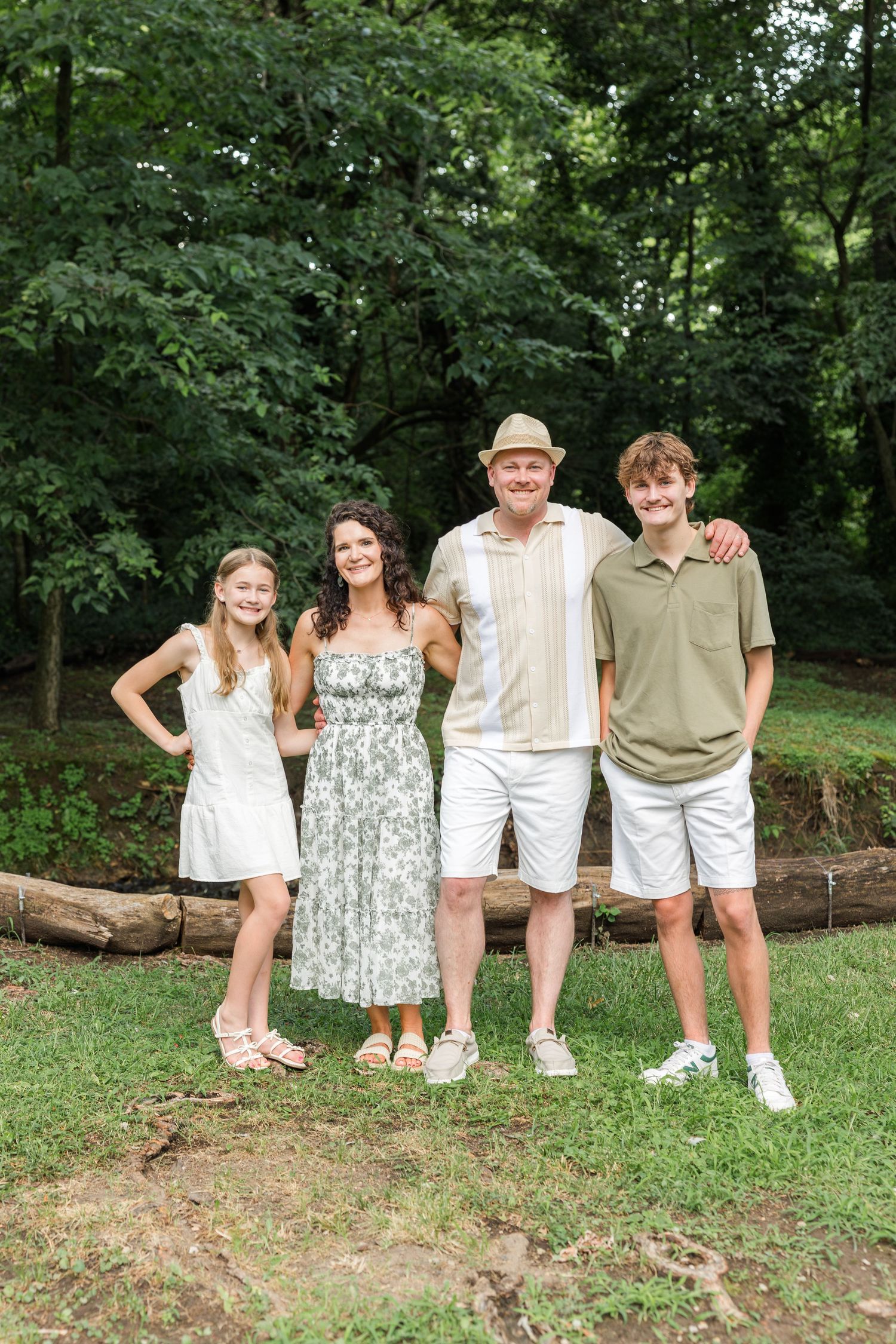 A group poses together outdoors on a grassy hillside surrounded by green trees in casual summer attire.