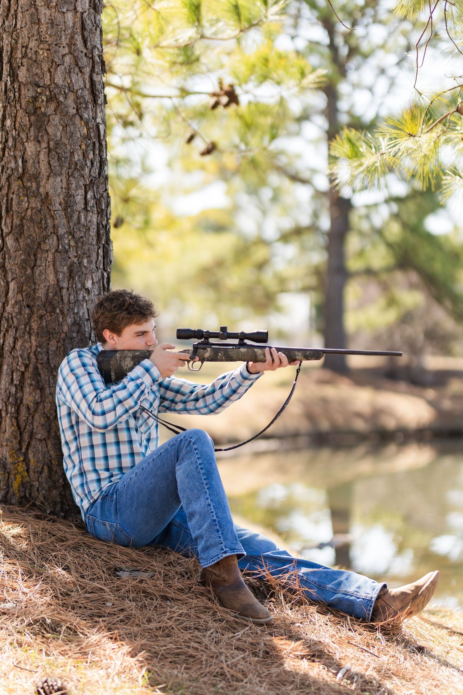 Person in plaid shirt and jeans aims rifle with scope while sitting against tree trunk near water.