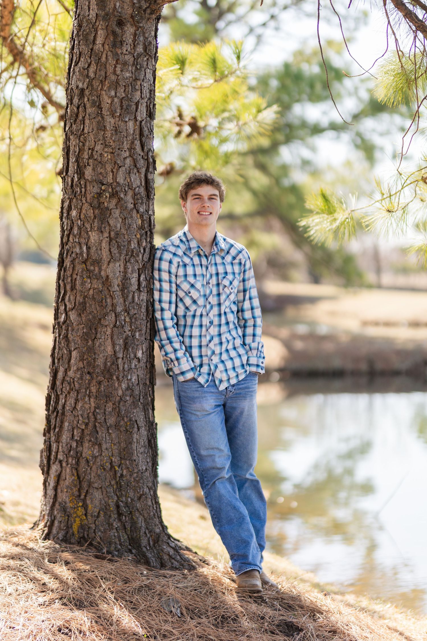 Person in blue plaid shirt leaning against pine tree near pond on sunny day.