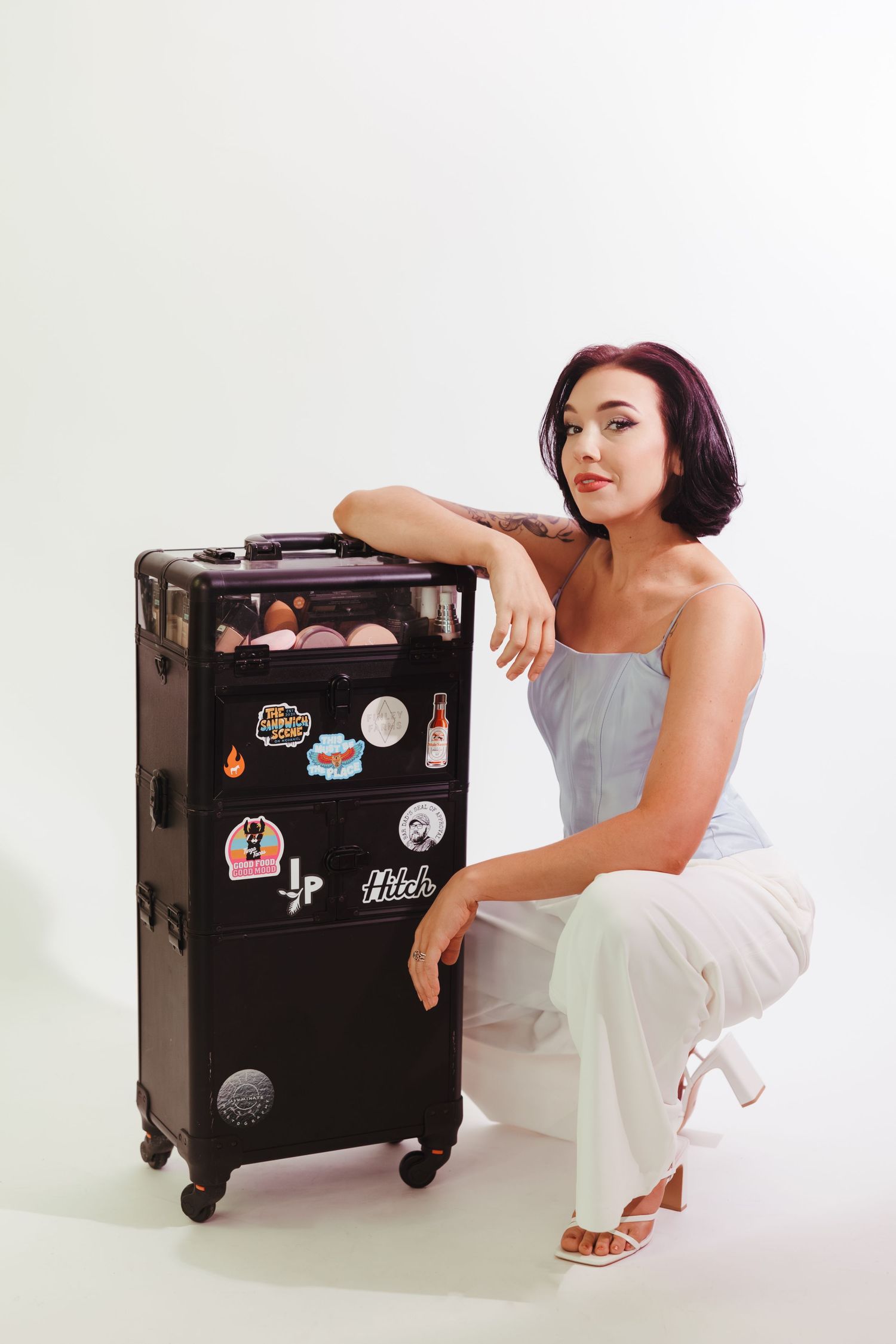 Person in white outfit poses with sticker-covered travel luggage against white background.