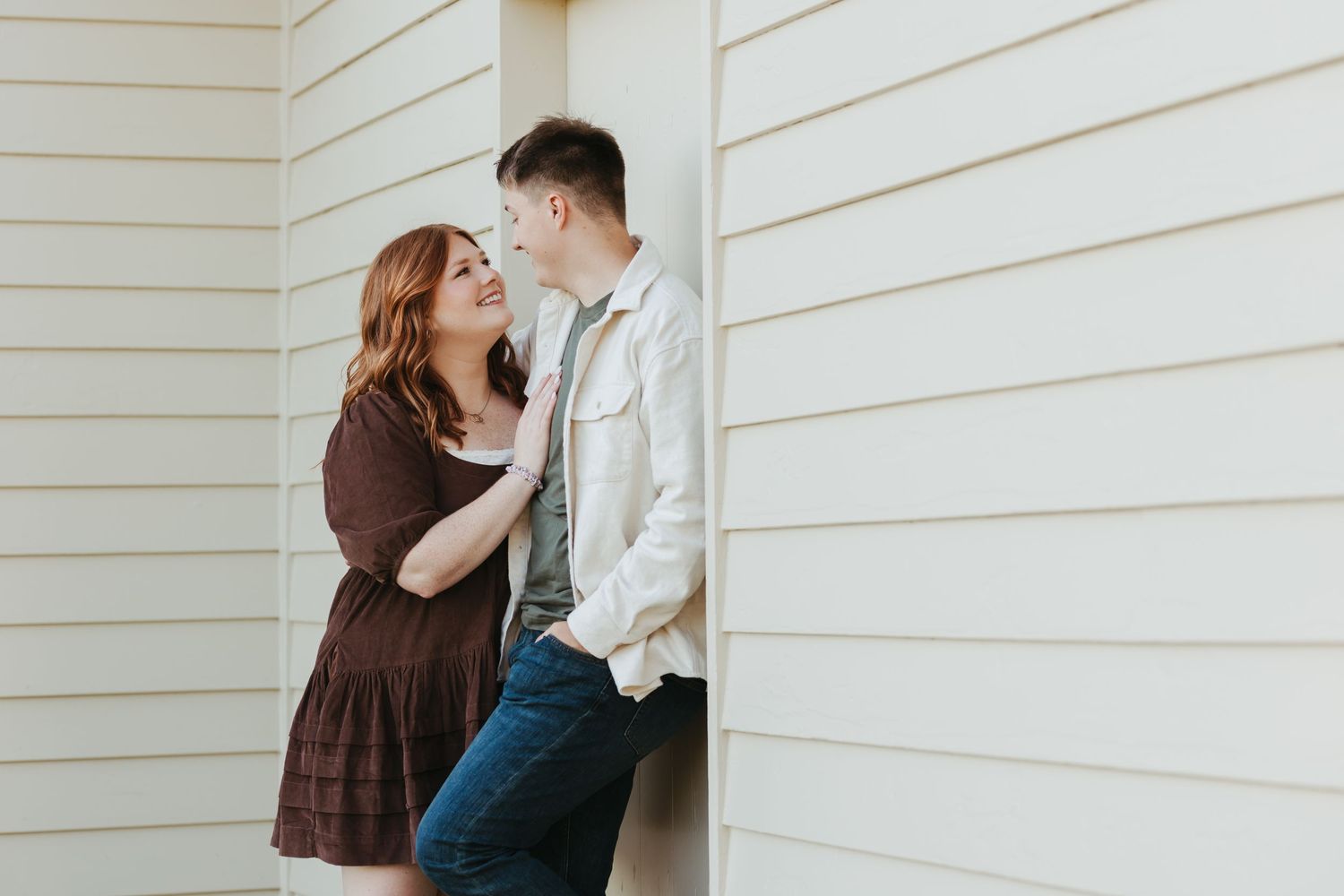 Romantic couple in brown dress and white jacket lean against exterior wall sharing tender moment.