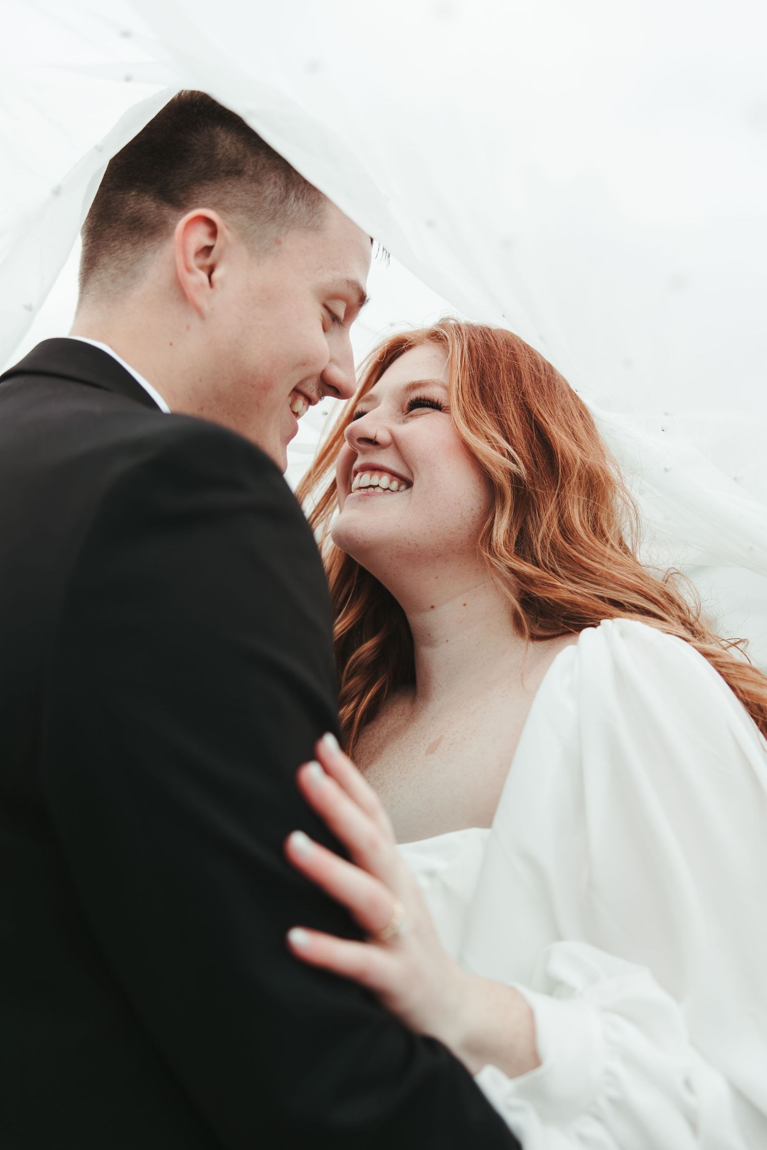 Close-up intimate moment between newlyweds showing joyful laughter under a wedding veil.