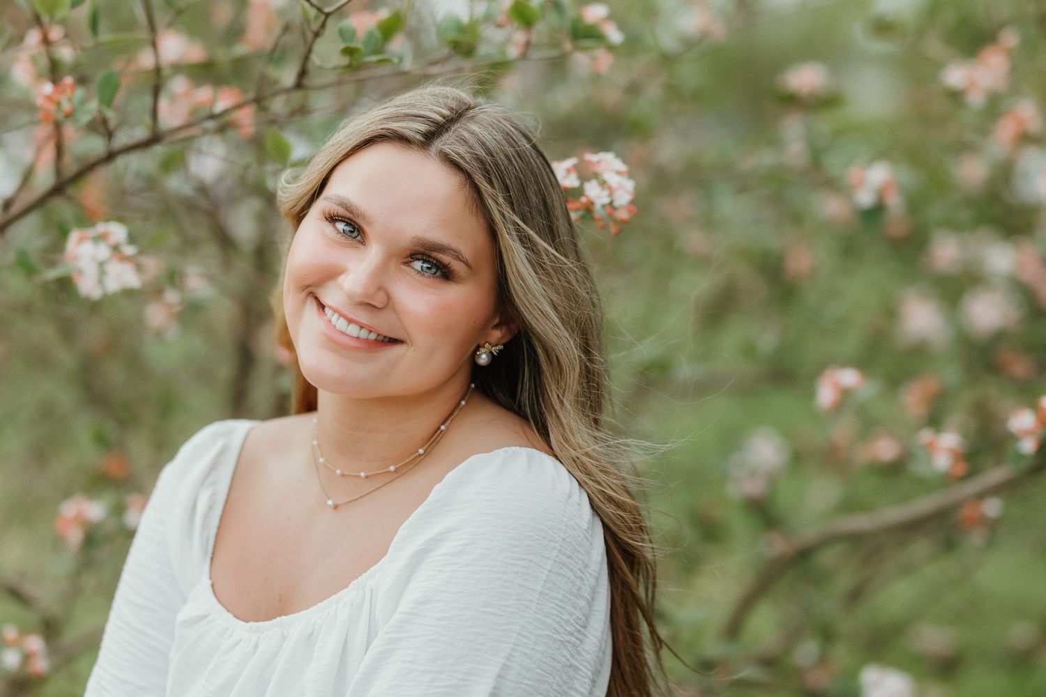 Portrait of someone in a white sweater smiling outdoors with pink blossoms in the background.