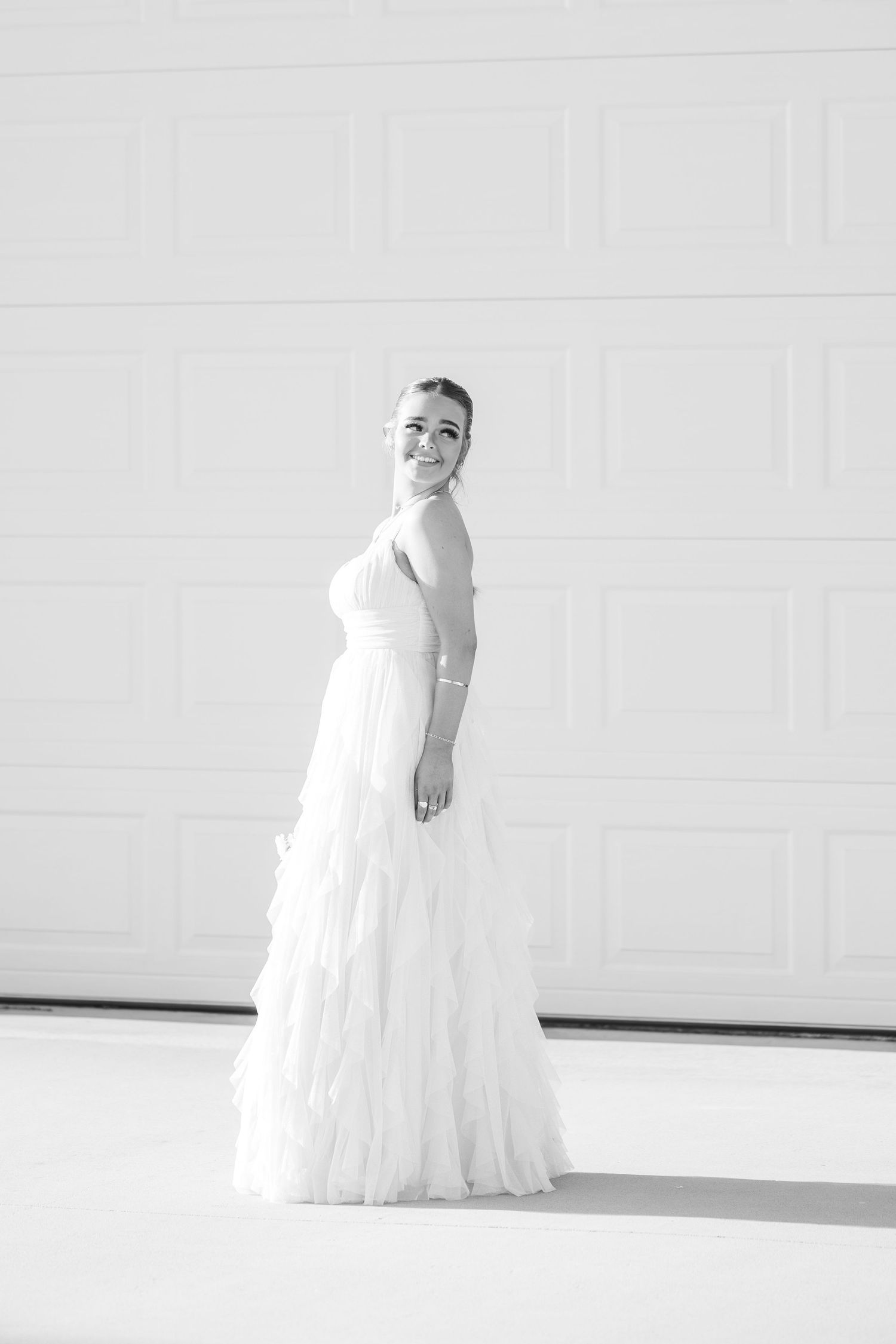 Black and white portrait of someone in an elegant white wedding gown with ruffled tiers standing against a light wall.