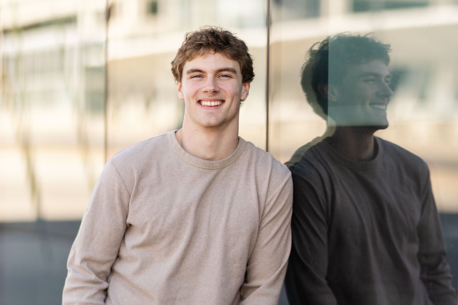 Person in beige sweater smiling while standing against reflective glass window creating mirror effect.