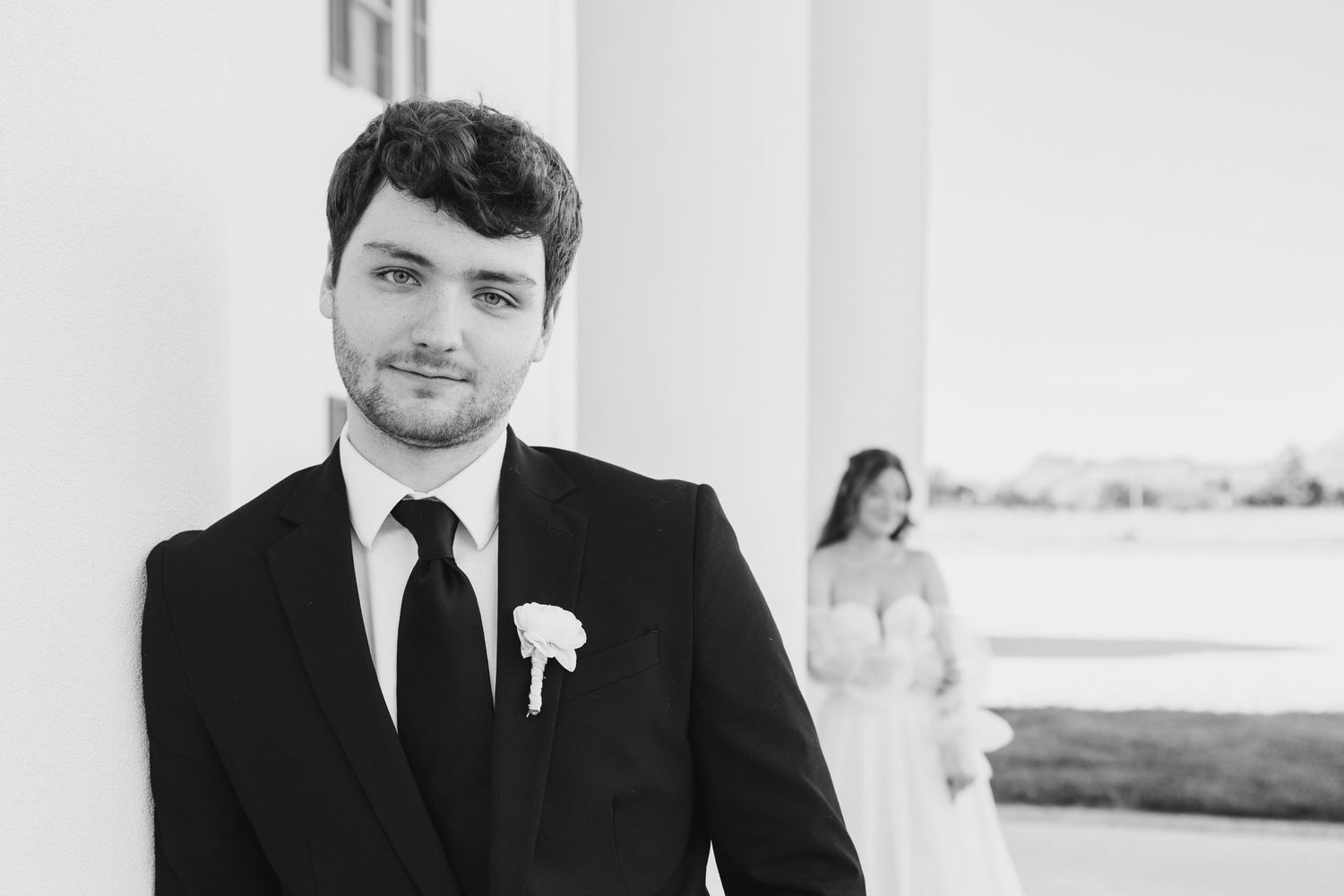 Black and white wedding photo of groom in suit with boutonniere, bride in white dress standing behind columns.