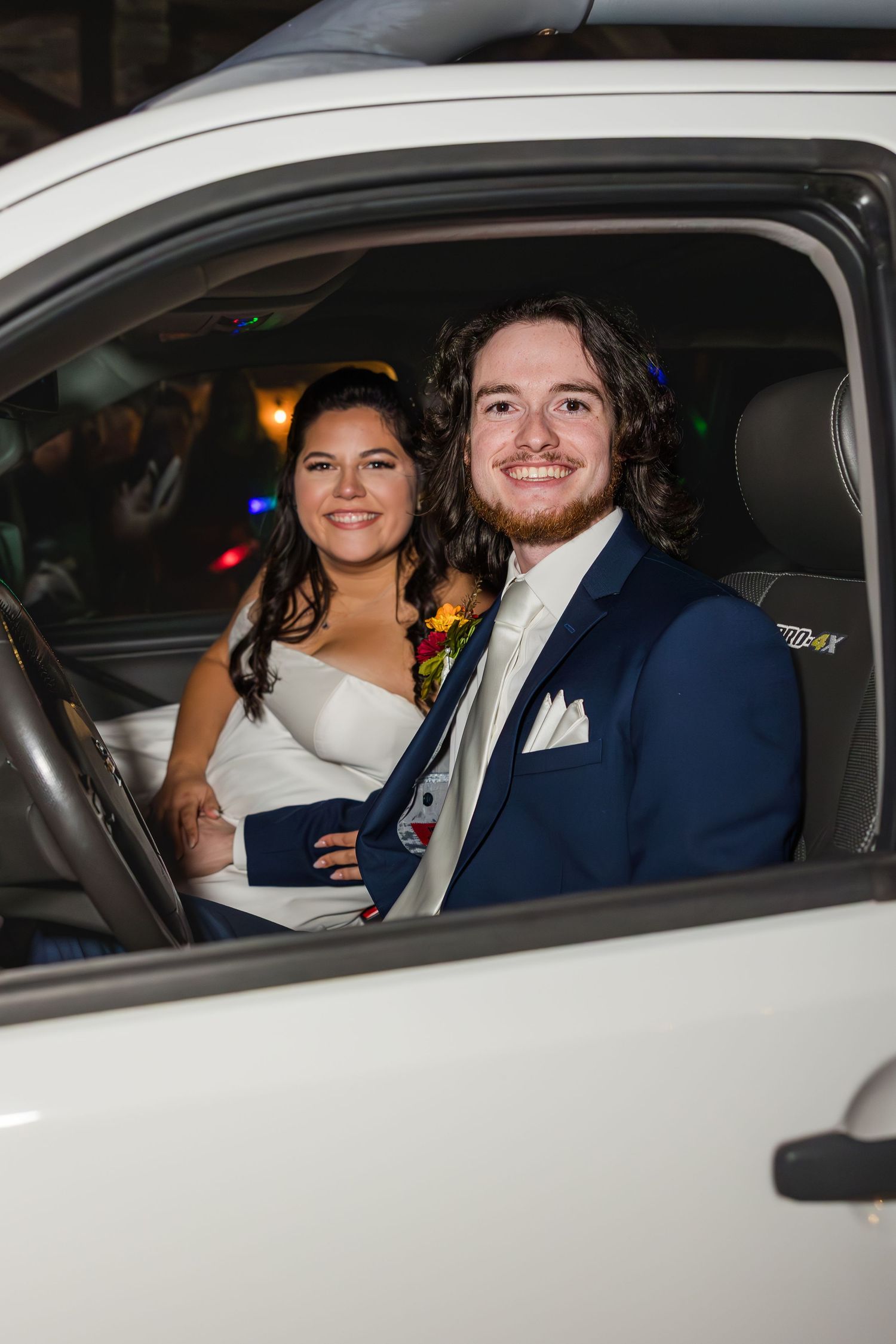 A smiling bride and groom in wedding attire sit together in a white vehicle on their wedding day.