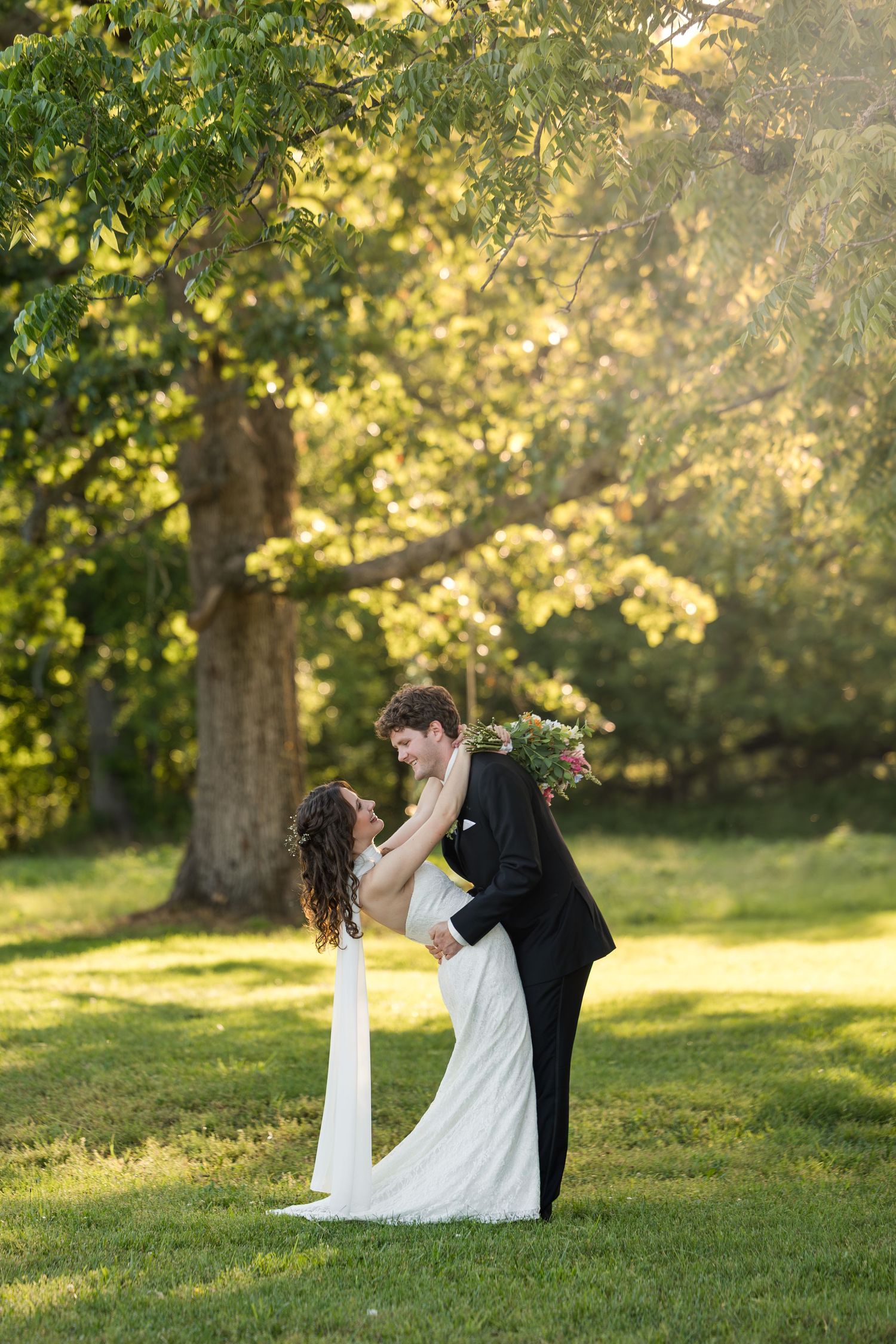 Groom lifting bride in white dress outdoors, surrounded by lush green trees in golden sunlight at a garden wedding.