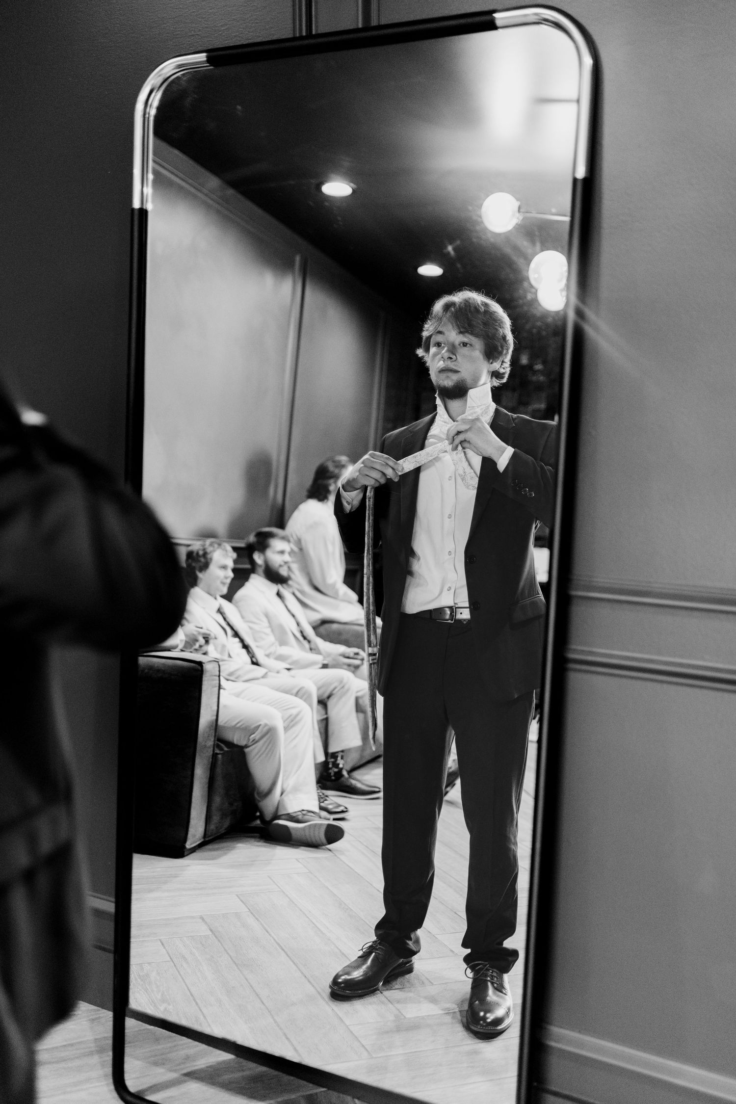 Groom adjusting bow tie while looking in mirror at wedding suit fitting, black and white photo.