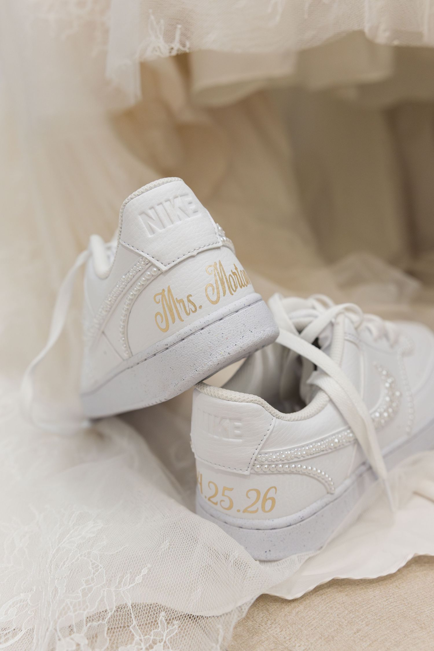 Custom white Nike sneakers with gold Mrs. and date 25.26 inscription displayed on wedding dress lace.