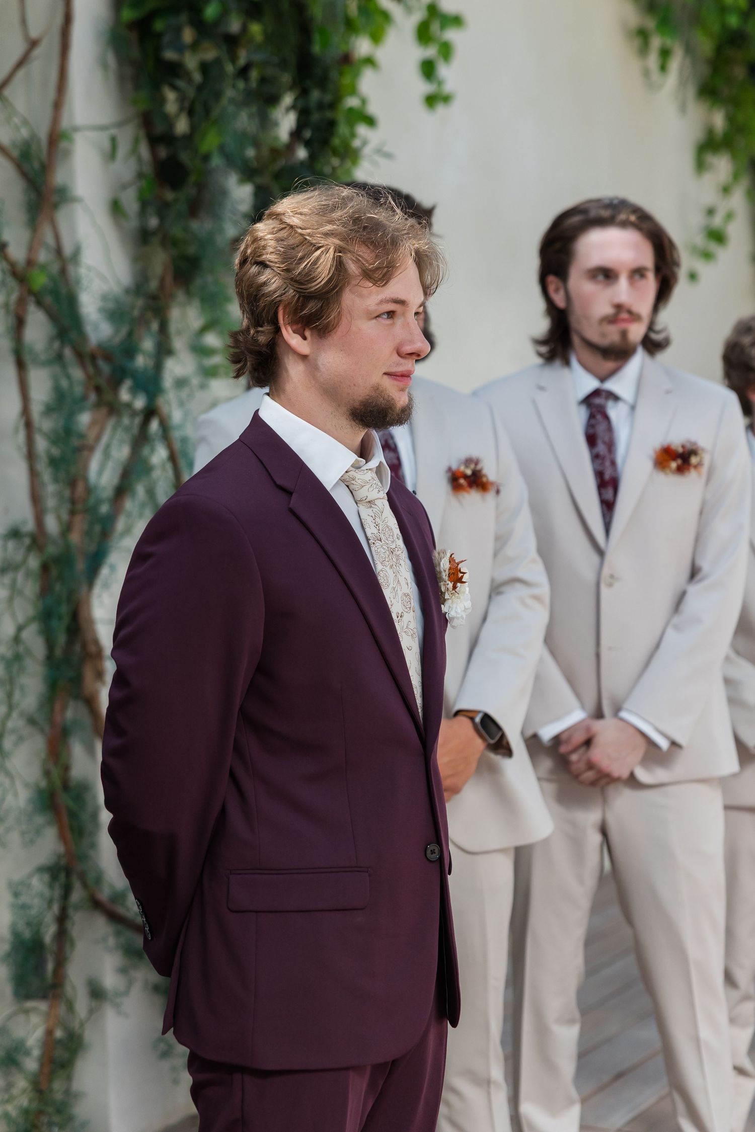 Groom in burgundy suit stands at altar with groomsmen in gray suits during outdoor wedding ceremony.