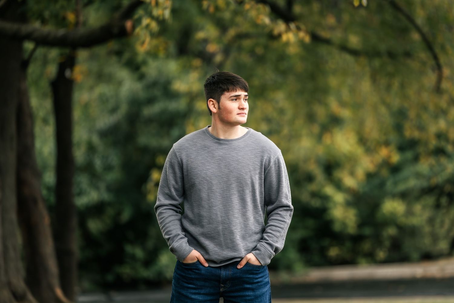 A person in a gray sweater and jeans stands casually against an autumn forest background.
