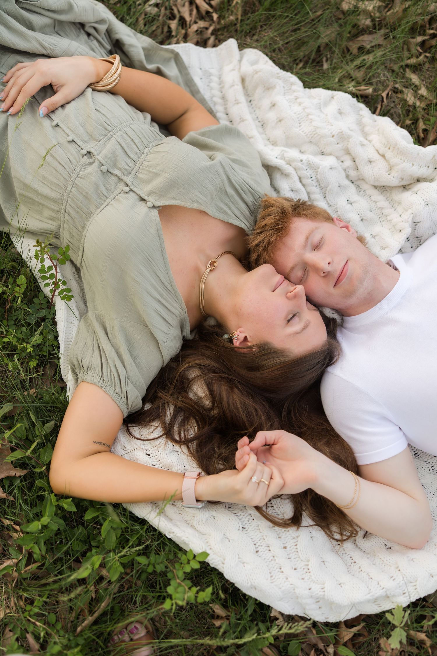 A couple lying together on a white blanket in the grass sharing a tender moment against a natural outdoor backdrop.