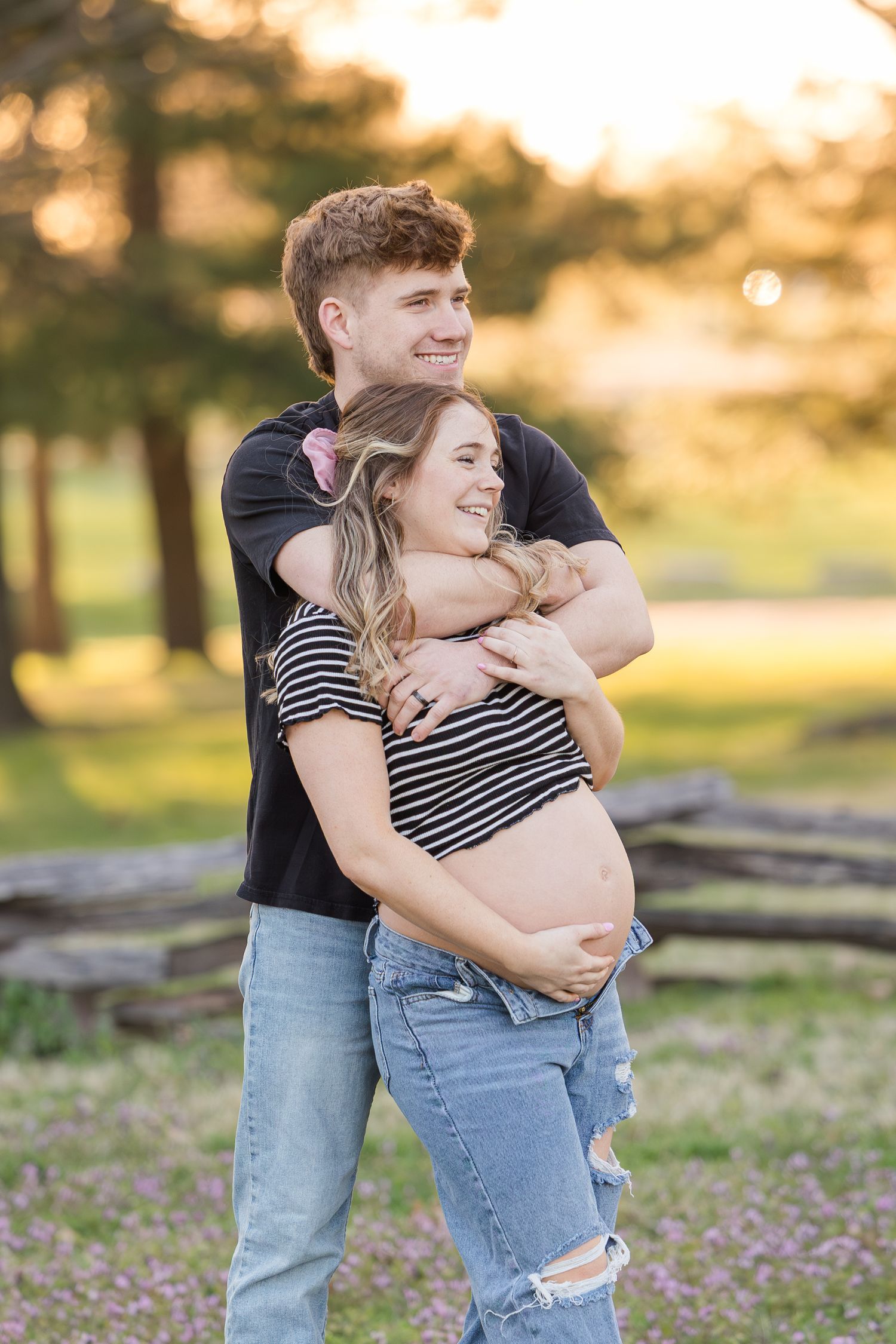 A couple embraces during sunset maternity photo session outdoors with warm golden lighting and trees in background.