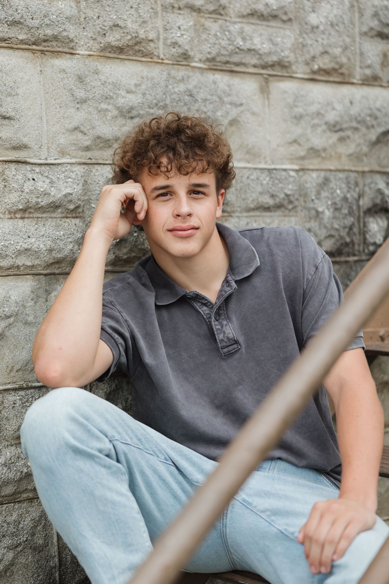 Young person in gray polo shirt and light jeans poses casually against stone wall outdoors.