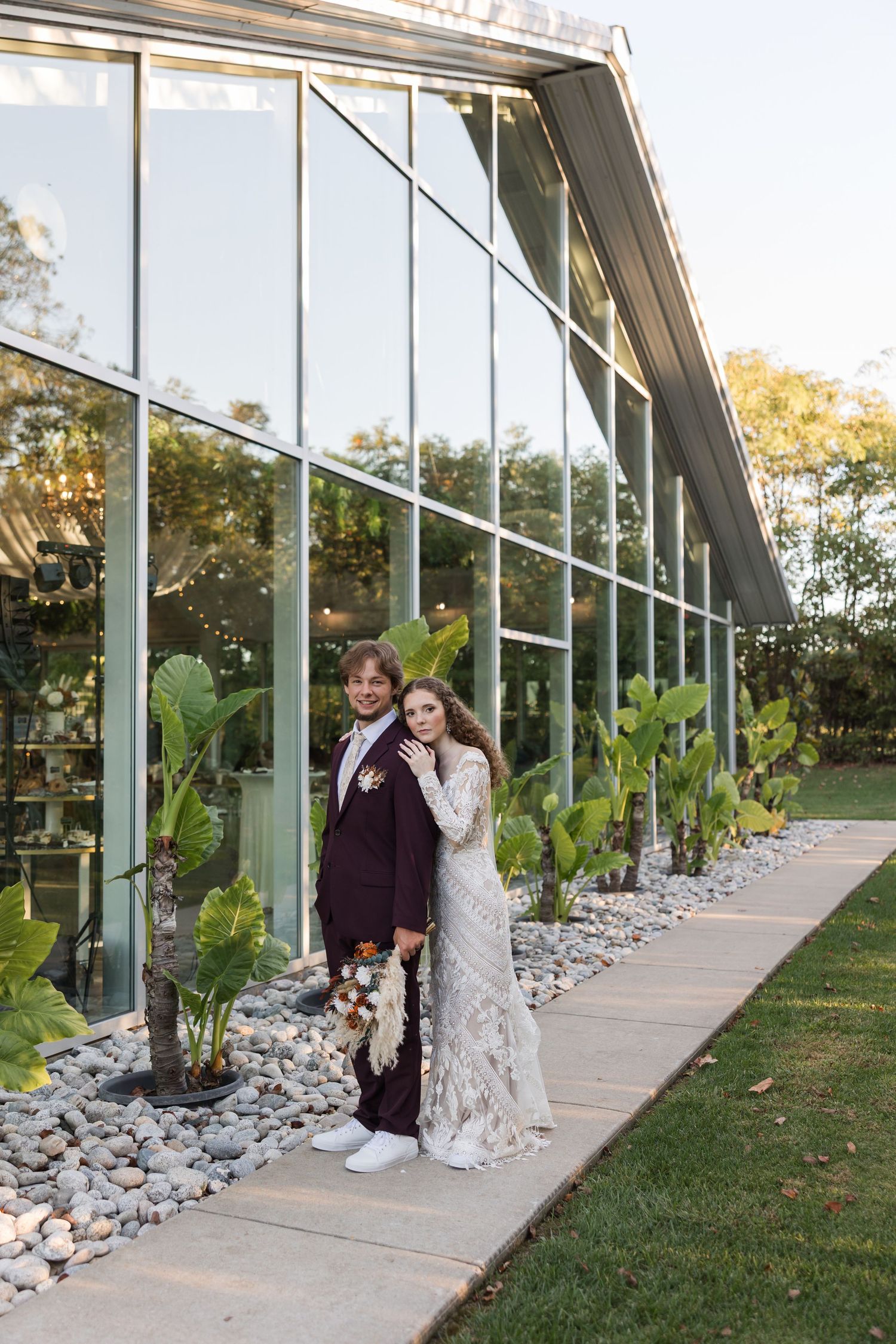 A couple poses together under a modern glass-walled walkway with landscaped rocks and greenery during golden hour.