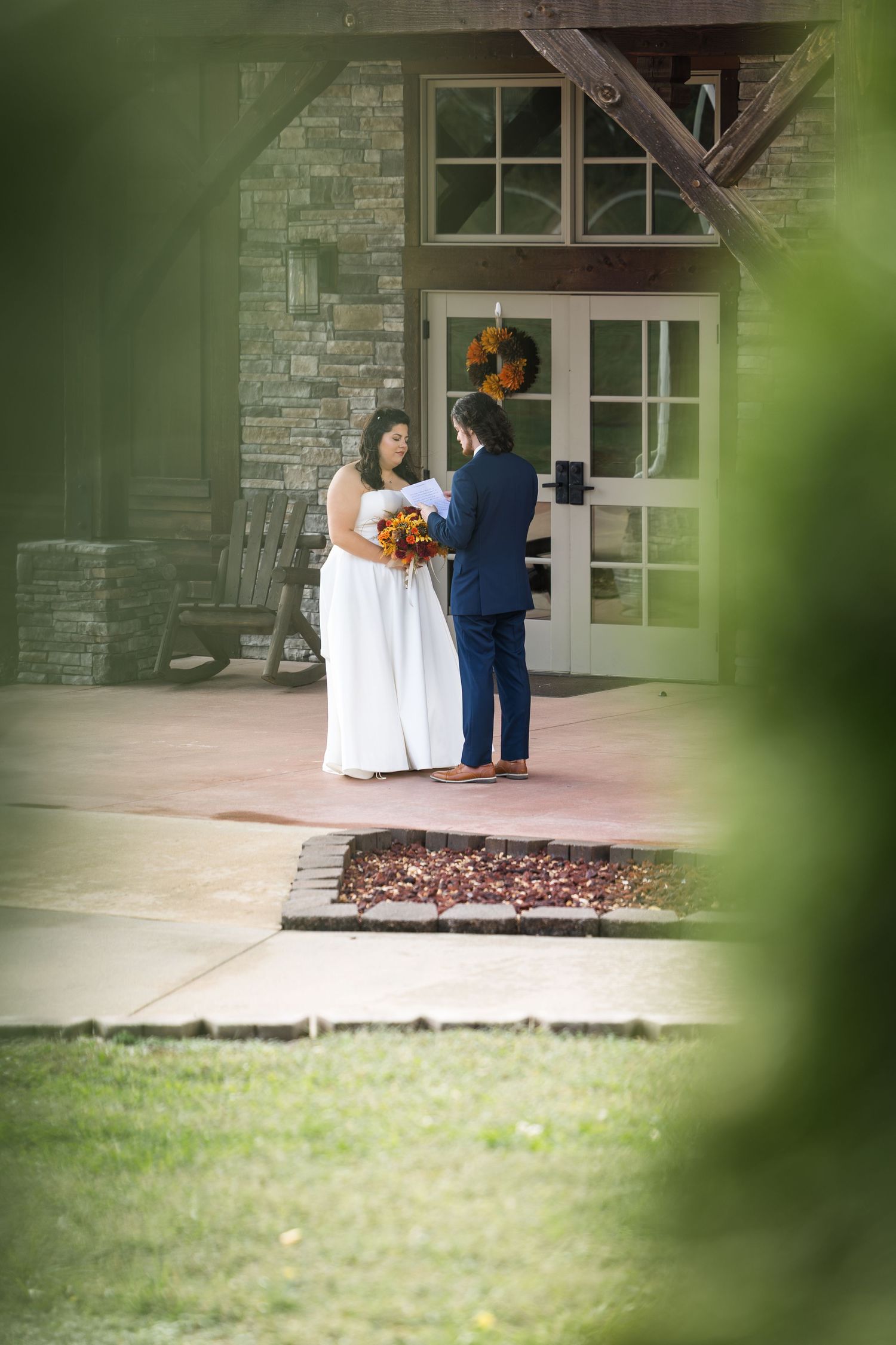 A wedding couple sits together on white chairs on a stone patio surrounded by greenery.