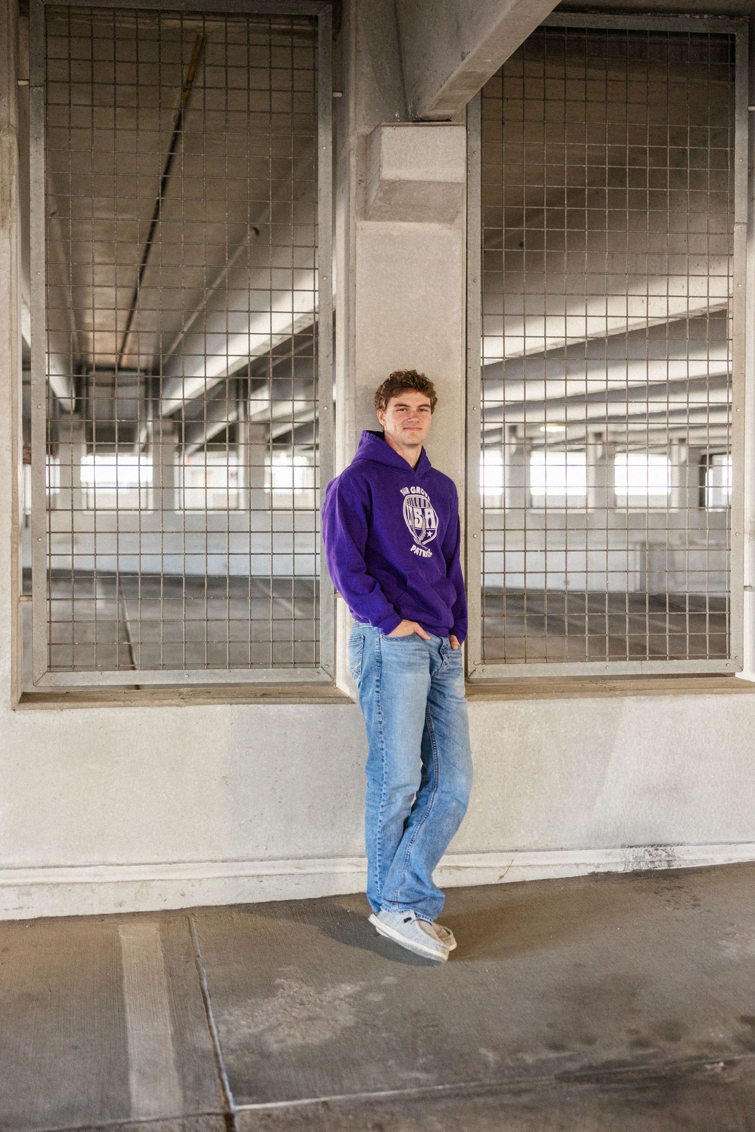 Figure in a purple hoodie and light wash jeans stands casually in an industrial concrete parking structure.